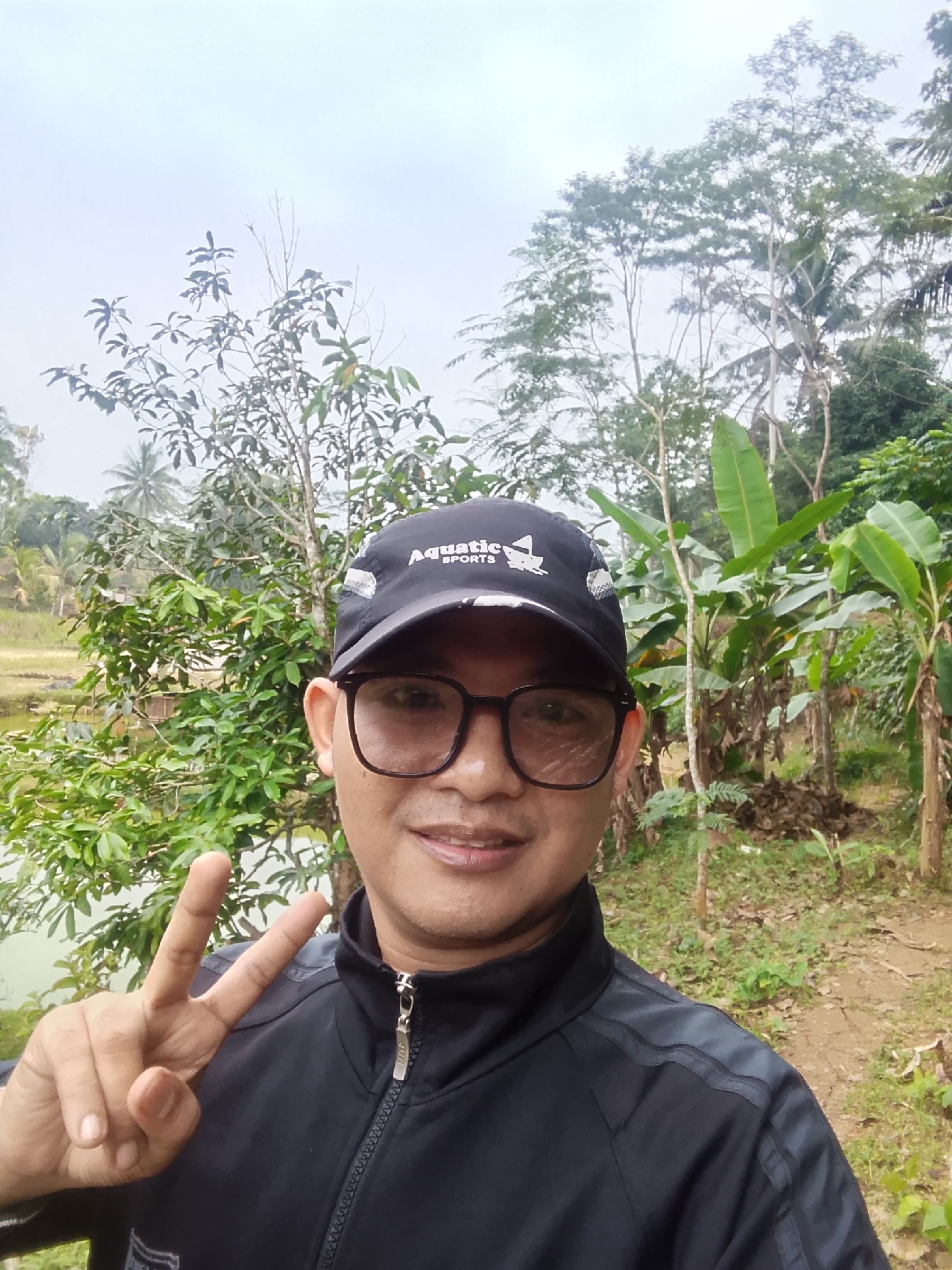 A man, wearing a black baseball cap with "Aquatic Sports" printed on it, and black-rimmed glasses, smiles at the camera. He is wearing a black jacket. His right hand is raised, making a peace sign. He is standing outdoors, with lush green trees and foliage behind him. The background also shows a body of water, likely a pond or lake, and what appears to be a rice field. The sky is overcast. The location is identified as Cikukulu, Indonesia.
