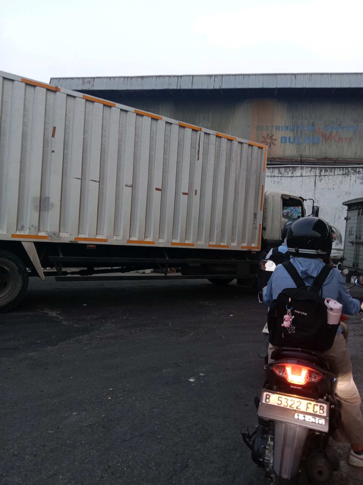 A low-angle, outdoor, eye-level, medium shot captures a scene on an asphalt road in Semarang, Indonesia. In the foreground, a person rides a black motorcycle, facing away from the camera. The rider wears a light blue hooded jacket, a black helmet with white accents, and tan pants. A black backpack with a pink bear charm and the word "merché" is attached to the rider's back. The motorcycle's license plate reads "B 5322 FCB" and "11-26". The motorcycle's taillight is illuminated red. To the left, the rear of a large, white, corrugated metal truck is visible, with orange reflective tape along its side. The truck's tires and lower chassis are also in view. In the background, a large, corrugated metal building with a darker roof extends across the frame. Faint blue lettering on the building reads "DISTRIBUTION CENTER BUILDING." The sky above is a pale, overcast gray.