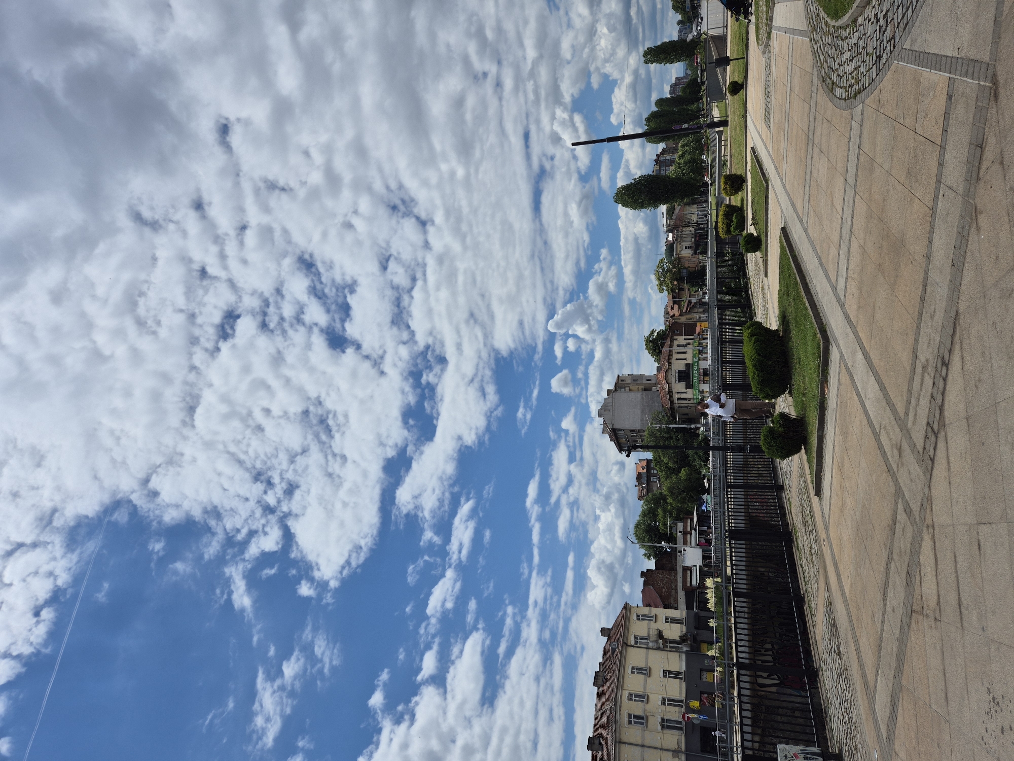The image shows a view of a riverbank in Sofia, Bulgaria, on a cloudy day. A man is standing on a paved walkway, facing the river. The walkway is lined with low green bushes. In the background, there are buildings and trees. A jet trail is visible in the sky. The river has a metal railing along its bank. The paving stones are a mix of square and rectangular shapes, with a cobbled border.