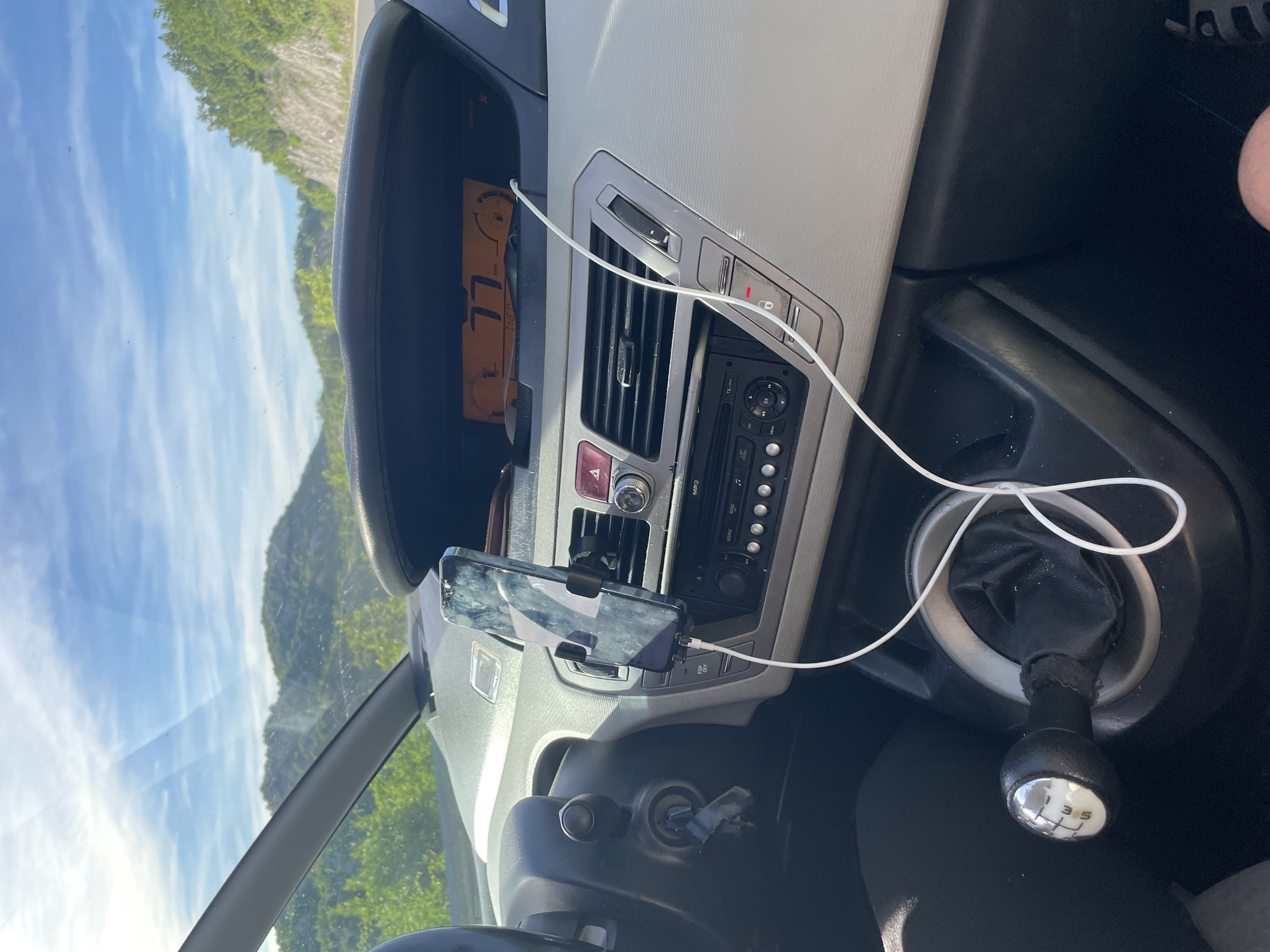 The image shows the interior of a car parked on a road in Očinići, Montenegro. The dashboard features a digital speedometer displaying "77" km/h, an air conditioning vent, and a car stereo. A smartphone is mounted on a holder on the dashboard, connected to a white charging cable. The car's gear shift, a 6-speed manual transmission, is visible in the foreground. The background reveals a sunlit, mountainous landscape with lush green vegetation. The sky is clear with scattered clouds.