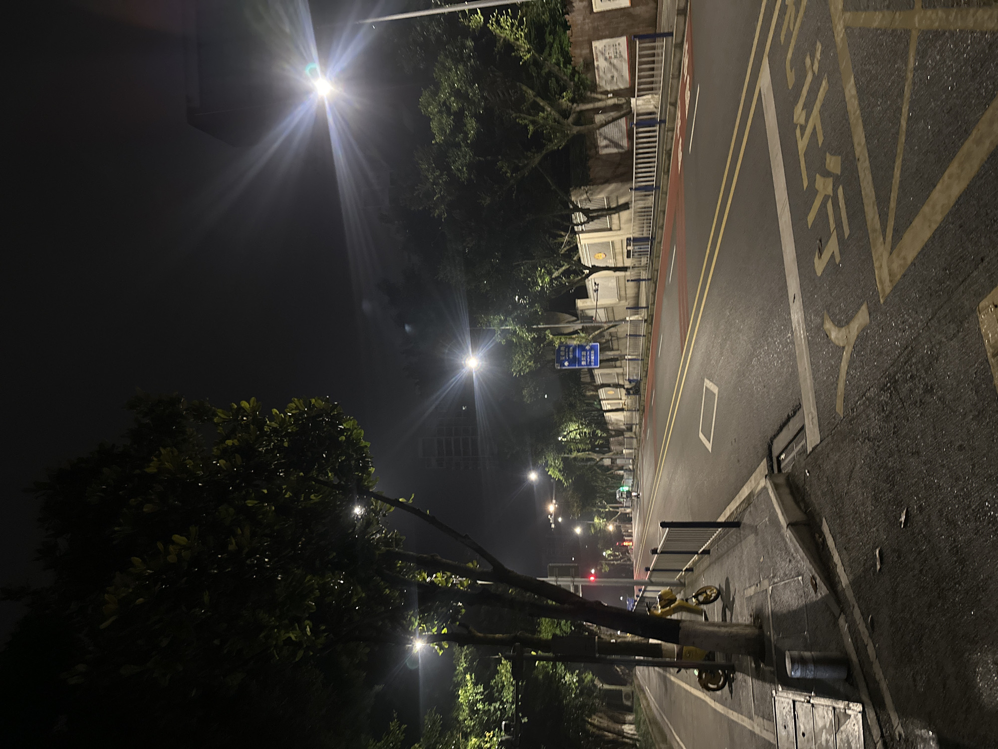 The image depicts an urban street scene at night in the Chongqing High-tech Zone, China. The street is illuminated by streetlights and the headlights of distant vehicles. Trees line the sidewalks, their leaves dark against the night sky. The road is paved and marked with yellow and white lines, indicating lanes and crosswalks. Some of the markings on the road are in Chinese characters. A few parked electric scooters are visible on the sidewalk. In the background, buildings and streetlights create a cityscape. The overall atmosphere is quiet and somewhat desolate, characteristic of a city at night.