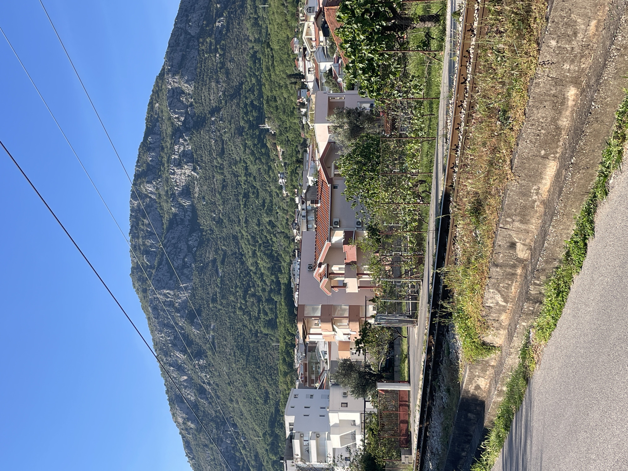 The image shows a hillside town nestled at the base of a large, rocky mountain covered in dense green vegetation. The scene is in the city of Bar, Montenegro. The sky is clear and blue. There are several buildings of varying heights and colors visible, mostly with white or beige facades and red-tiled roofs. A series of power lines run across the upper left of the frame. Along the right side of the image, there is a road and a concrete barrier or ditch, with vegetation growing within and along it. There are also fences and some trees with dense foliage closer to the buildings. The angle of the image is such that the mountain dominates the left side, while the town stretches along the base towards the right.