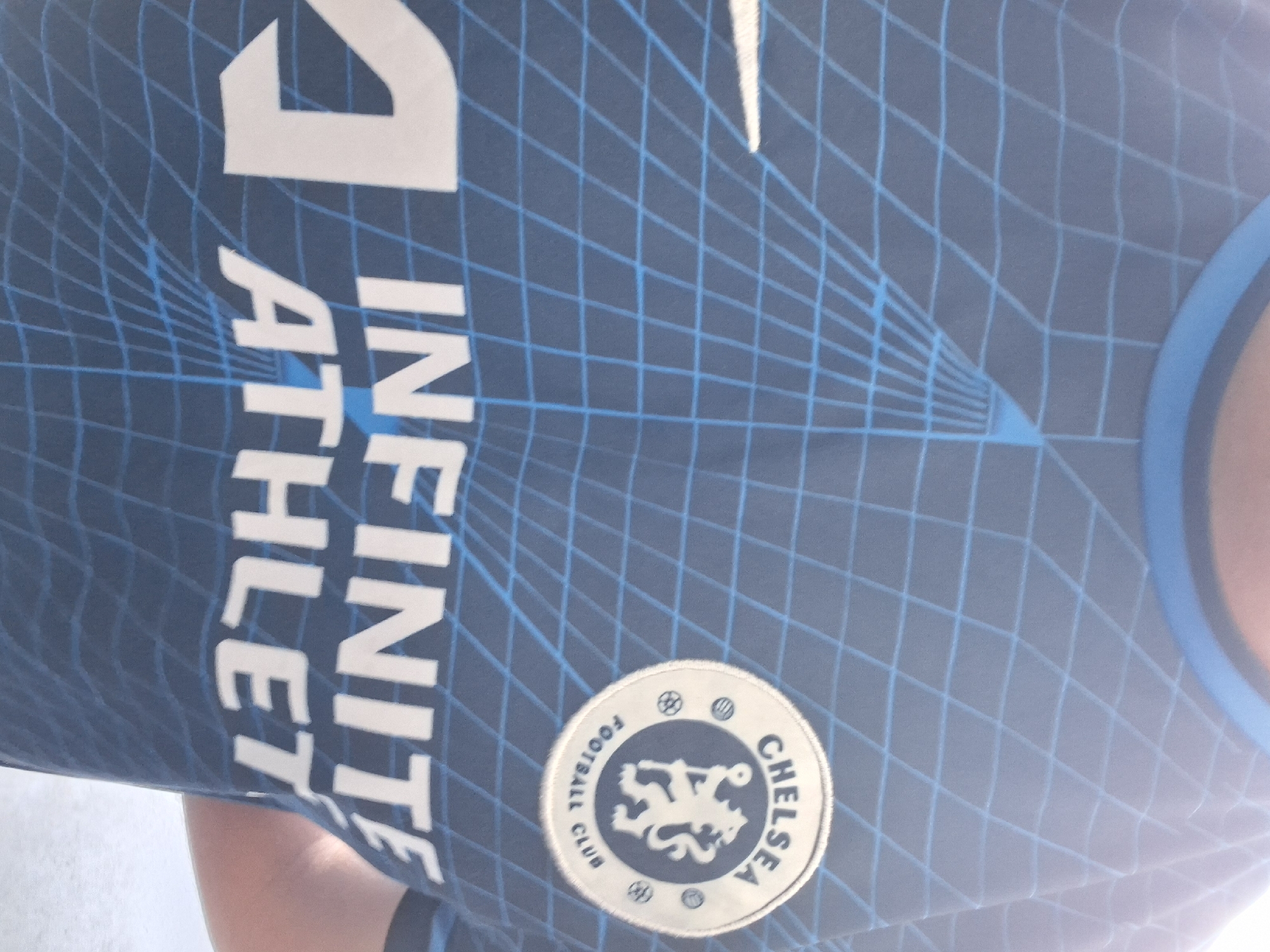 A close-up image shows a Chelsea football club jersey. The jersey is predominantly blue with a light blue grid pattern overlaying the fabric. The Chelsea club badge is visible on the lower right side of the jersey. To the left of the badge are the words "INFINITE ATHLETE" stacked vertically in white letters. Above the words is a white swoosh symbol, typical of the Nike brand. The jersey has a blue collar trim.