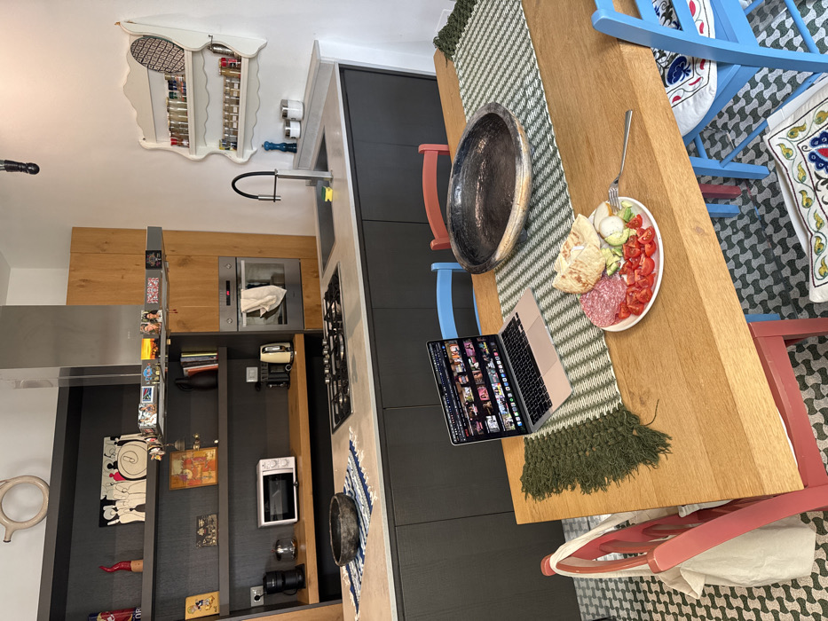The image displays a modern kitchen and dining area located within a residence in Lecce, Italy. A rectangular wooden dining table occupies the foreground and midground, covered by a green and white striped table runner. On the table, a silver-colored laptop is open, displaying an interface with numerous video thumbnails. Adjacent to the laptop, a large, dark, metallic-finished bowl rests on the runner. A white plate positioned to the right holds a meal consisting of sliced salami, chopped red tomatoes, sliced cucumber, a portion of white cheese or burrata, and pieces of flatbread, accompanied by a silver fork. Several chairs with contrasting colors (red and blue) surround the table, some featuring patterned cushions, and a light-colored canvas bag hangs from one red chair. The floor is tiled with a geometric pattern in dark gray and off-white.
The background reveals a contemporary kitchen area. Dark gray base cabinets support a light gray countertop, which incorporates a gas stovetop, a rectangular sink with a black gooseneck faucet, and a built-in oven with a white cloth hanging from its handle. Overhead, a stainless steel range hood is visible. To the left, a section of dark gray open shelving with wooden accents holds various decorative items, including artwork, books, and small figures. Appliances such as a microwave, coffee maker, and electric kettle are present on a wooden shelf segment. On the right wall, a white, ornate two-tiered shelf is mounted, filled with spice jars.