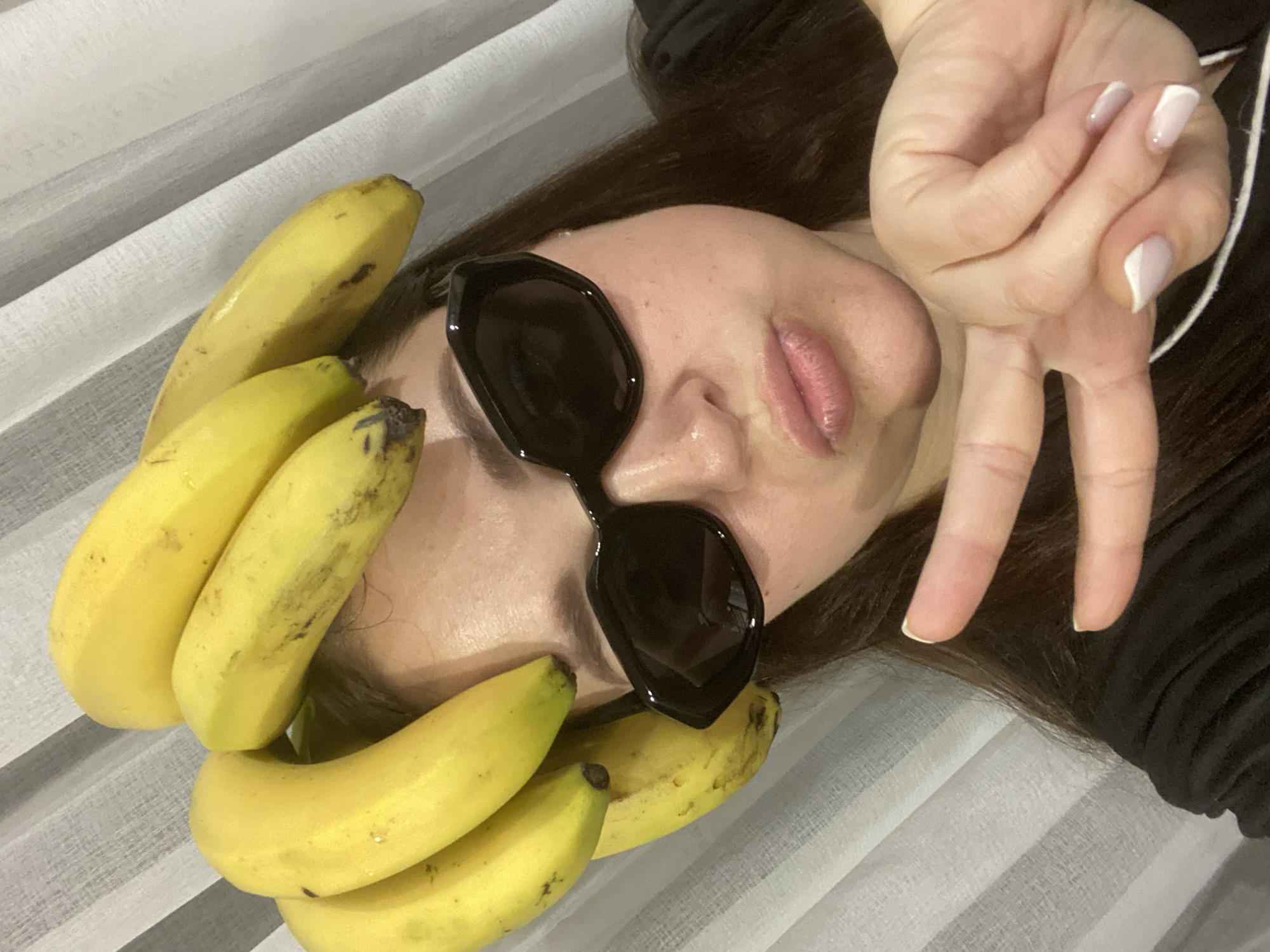 An overhead, slightly angled shot shows a person (likely a woman) lying down, with their head resting on a surface. Their long dark hair spreads out around their head. A bunch of ripe yellow bananas, some with small dark spots, is whimsically arranged around the person's face, partially covering their hair and neck.
The person is wearing stylish black sunglasses with an octagonal shape, obscuring their eyes. Their lips are slightly parted, revealing a natural pink hue, possibly enhanced with lip gloss or a subtle lipstick. One hand is raised and positioned near their head, forming a "peace" sign with the index and middle fingers extended. The nails on this hand are neatly manicured with a light, possibly nude or pale pink, base and white tips.
The background consists of a light-colored, striped fabric, which could be curtains, bedsheets, or a textured wall covering. The overall mood appears playful and relaxed. There is no visible text in the image. This scene takes place indoors, in Sochi, Russia.