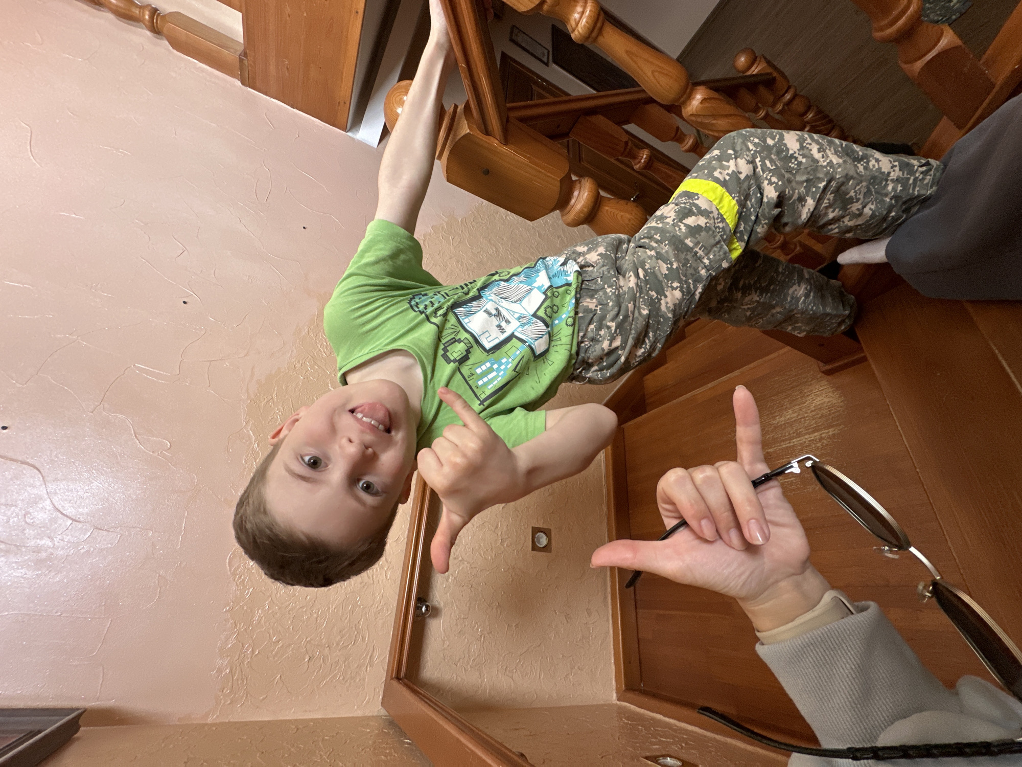 A young boy is hanging upside down from a wooden staircase railing in an indoor environment. He is wearing a green t-shirt and camouflage pants with a yellow stripe. He is smiling, sticking his tongue out, and making a hand gesture with his right hand. A person's arm is visible in the foreground, also making a hand gesture and holding a pair of sunglasses. The walls are light peach, and the floor appears to be carpeted. The scene is likely indoors, possibly inside a residence. Ilyinsky, Russia is not discernible from the image.