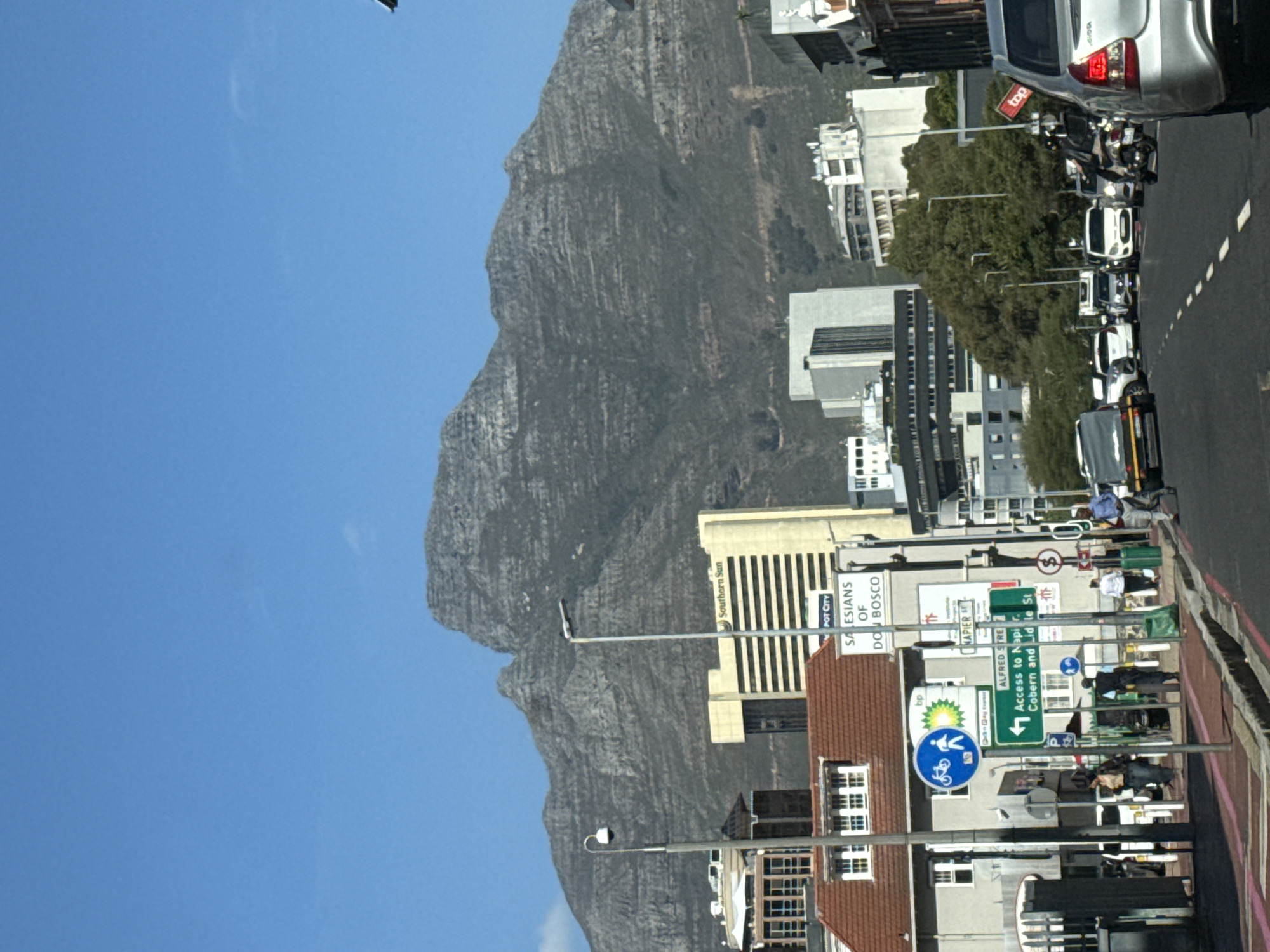 The image, taken in Cape Town, South Africa, captures a sunny daytime street scene with Table Mountain dominating the background under a clear blue sky. The photo is oriented vertically, showing the mountain's rugged, grey, and partially vegetated slopes rising behind an urban area.
In the foreground and midground, a bustling city street is visible. Buildings line the road, including a modern, light-colored high-rise with vertical elements, identified by the text "Southern Sun" and "POT CITY". Adjacent to it is a building with "SALESIANS OF DON BOSCO" written on its facade. A red-tiled roof building is also prominent.
The street features several parked cars, including a silver Toyota SUV at the upper right and a white pick-up truck further down. Pedestrians are visible on the sidewalks, with some individuals standing or resting. Street lamps and various traffic signs are interspersed throughout the scene. Green trees line parts of the street, adding foliage to the urban landscape.
Notable signs include: "NAPIER ST", "ALFRED STRE" (likely "Alfred Street"), and a green directional sign indicating "Access to Napier, Cobern and Lid le St". A "bp" (BP) logo and a blue circular sign with a bicycle symbol are also discernible. The overall atmosphere suggests a typical day in a vibrant city, with the iconic Table Mountain as a majestic backdrop.