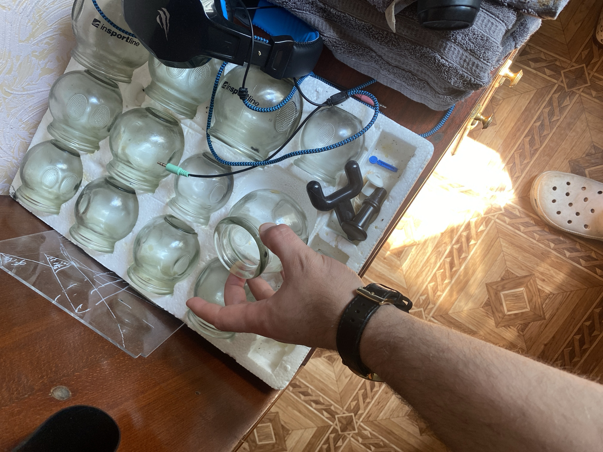 This is a top-down, eye-level shot of a collection of glass massage cups and a massage tool laid out on a white foam insert, which rests on a wooden surface. A person's arm and hand, wearing a dark leather watch, are reaching into the frame, holding one of the glass cups. In the background, headphones with a blue cord are visible, as well as a grey towel. The scene is brightly lit, suggesting daytime. The floor has a wood-grain pattern with geometric inlays. Visible text on the glass cups reads "insportline". A white shoe is present in the lower right corner. The overall impression is one of preparation for a massage or therapeutic treatment.