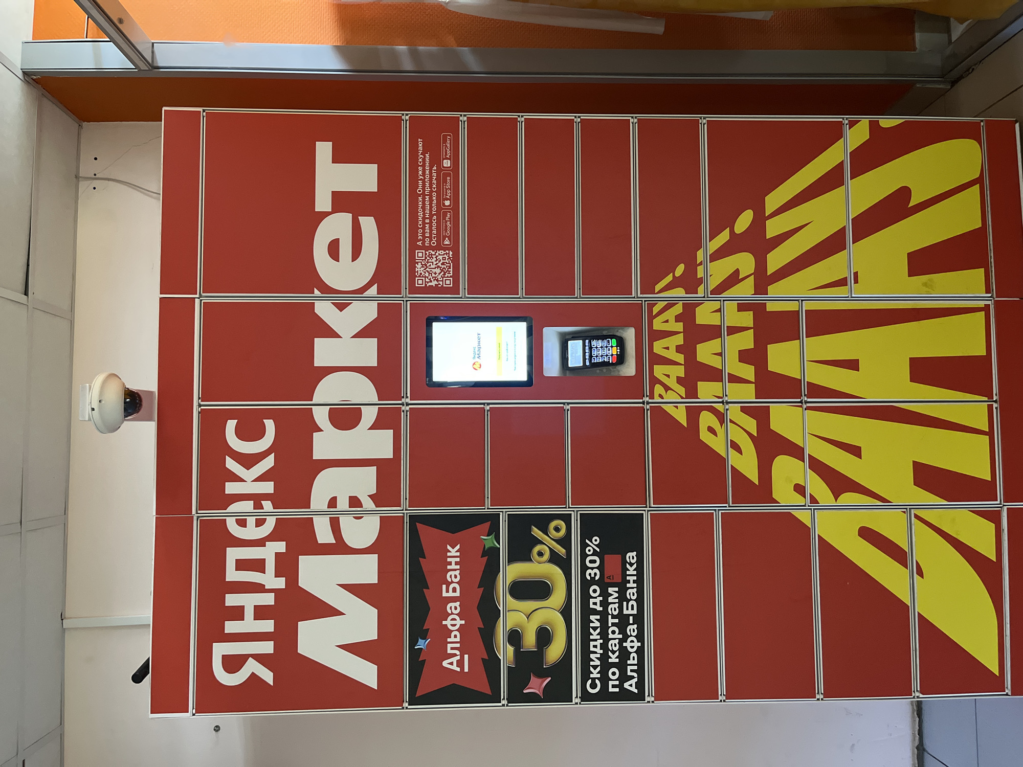 This is an indoor shot of a Yandex Market automated parcel locker in Klin, Russia. The locker is a red cabinet with a white metal frame. The left side displays the "Yandex Market" logo in Russian. Below that is an advertisement for Alpha Bank offering up to 30% discounts on purchases. The right side of the cabinet is covered in large yellow lettering. In the center, there is a screen and a card reader. At the top left of the image, there is a white security camera mounted on the wall. Above the cabinet is an orange wall or ceiling.