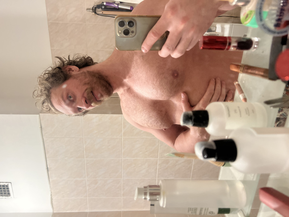 A muscular man takes a selfie in a bathroom mirror. The shirtless man holds his phone, displaying a three-camera array. He has curly blond hair, a beard, and fair skin. The reflection reveals toiletries and other items cluttered on the bathroom counter. The background shows white tiled walls and some hanging device.