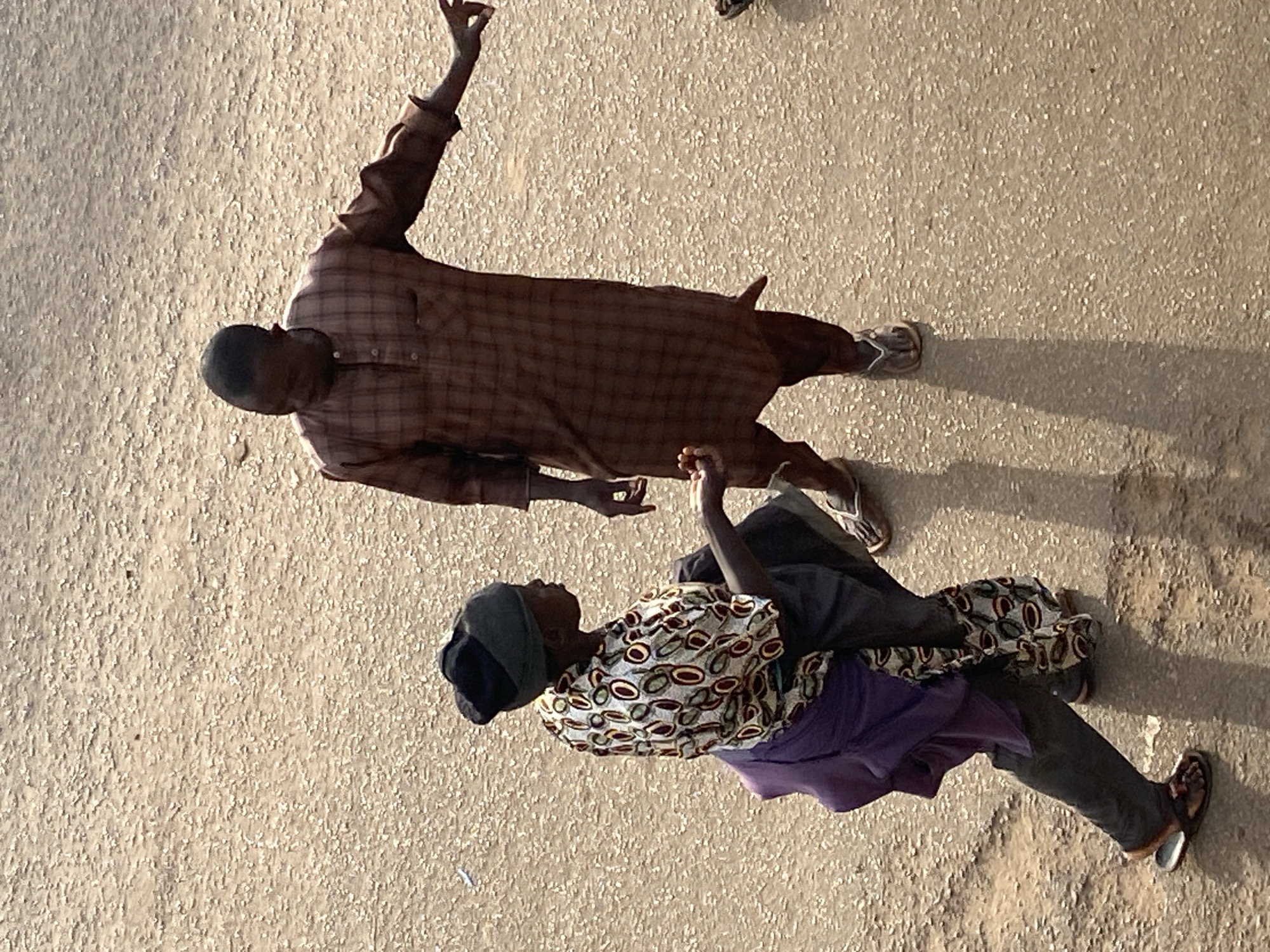 In Bukuru, Nigeria, two individuals are observed standing on a dirt road. A person on the left, wearing a patterned shirt and dark trousers, is kneeling and appears to be in conversation with a person on the right. The individual on the right is wearing a plaid shirt and trousers and is gesturing with their right hand. The ground is dry and textured, with a visible shadow cast by the person on the right.