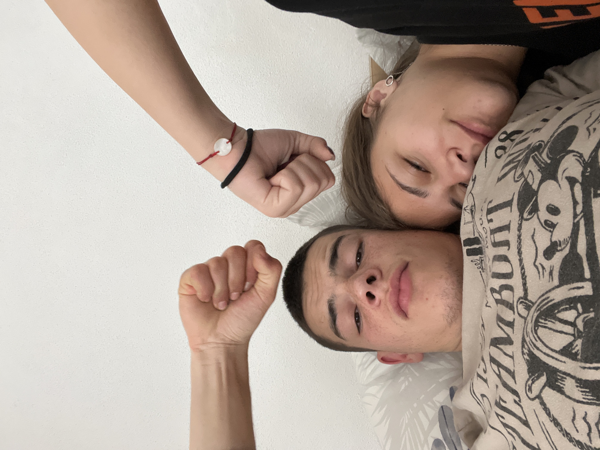 A selfie shows two people lying down with their fists raised. A young woman is lying on her side, with her face visible, wearing a black top and silver hoop earring. A young man is next to her, also lying down, with a white patterned shirt. Two arms are visible, both with clenched fists raised upwards. The woman has a bracelet on her wrist with a white round disc and a red string. They are indoors against a white wall. The exact location within Karavelovo, Bulgaria, is not discernible from the image.