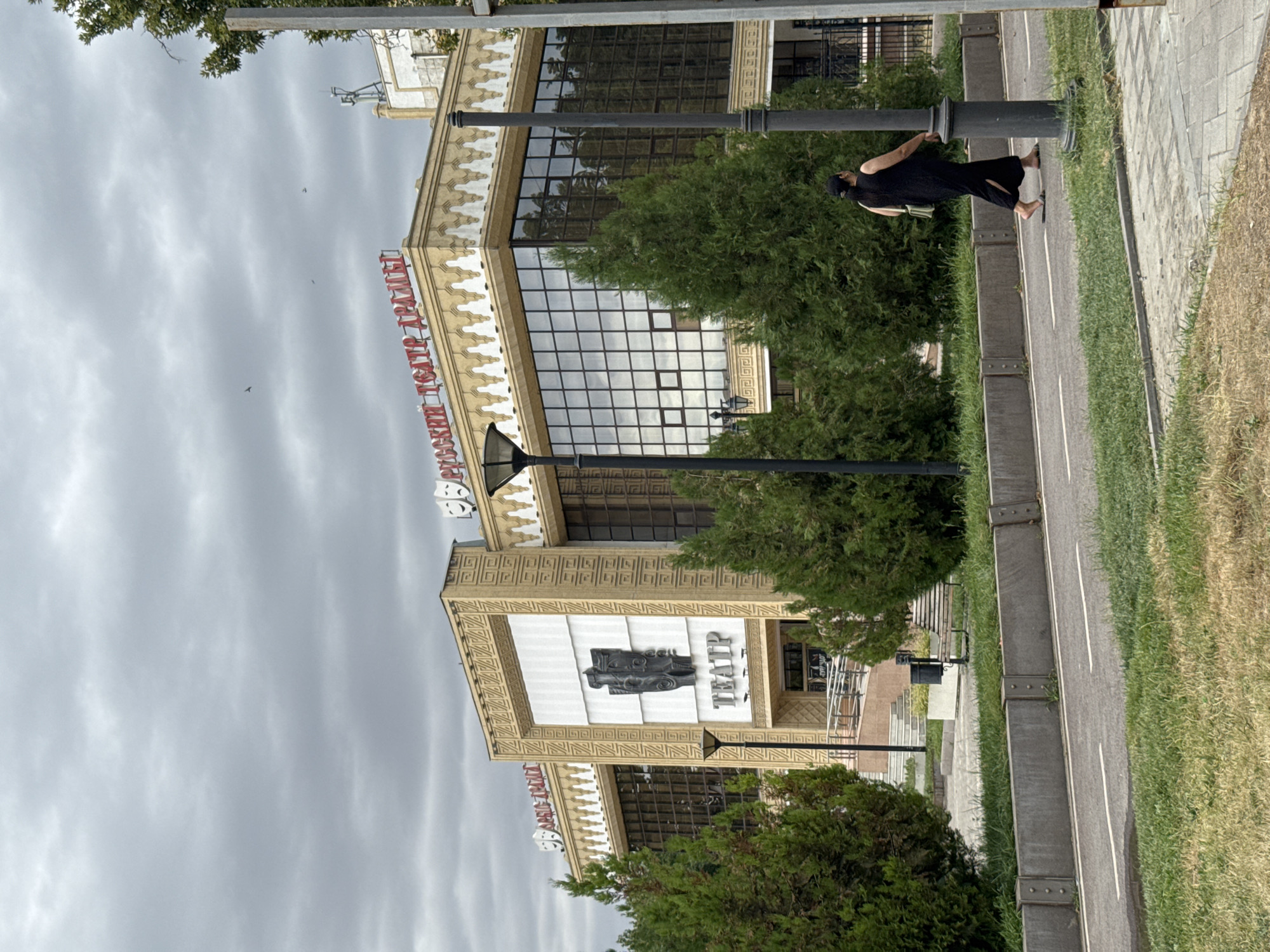 A woman in a long black dress and flip-flops walks on a paved path in front of a theater building in Shymkent, Kazakhstan. The theater, identified by red Cyrillic lettering reading "РУССКИЙ ТЕАТР ДРАМЫ" (Russian Drama Theater), has a facade with decorative carvings and large windows. Green trees are visible on both sides of the building and in the foreground. The sky is overcast with clouds, and there are some birds flying in the distance. A lamppost stands to the right of the woman.