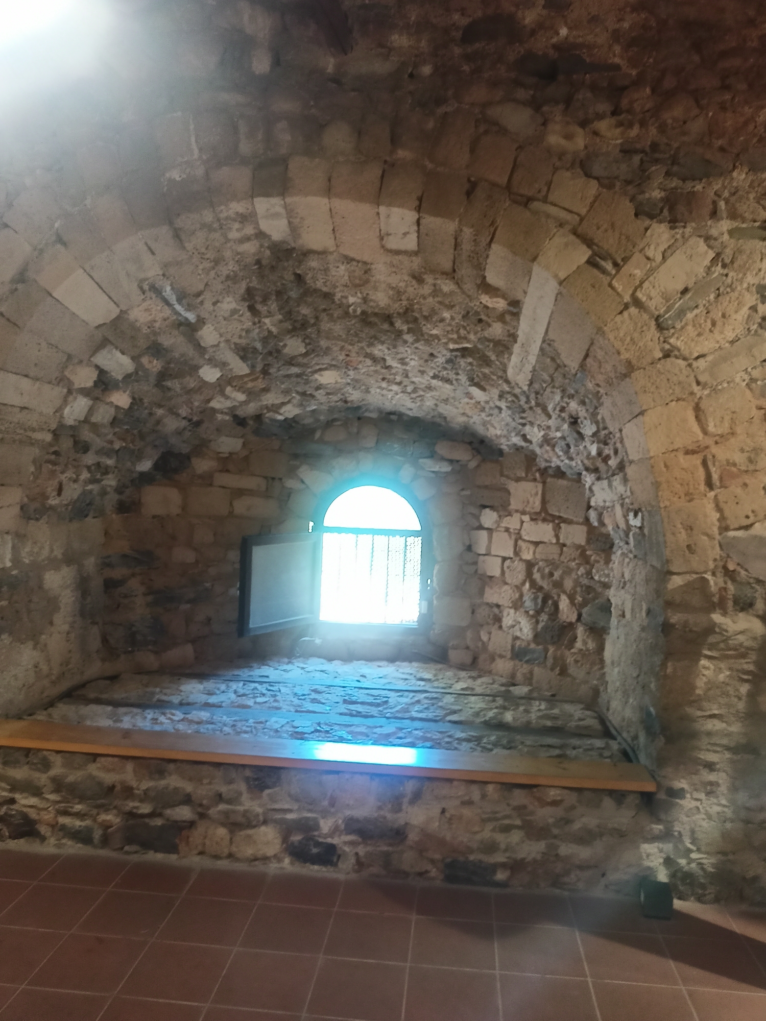 The image depicts the interior of a stone structure with a vaulted ceiling and rough stone walls. A semicircular window, partially open, is visible in the far wall, allowing bright light to illuminate the area. The window has a metal grate. In front of the window is a wooden bench or ledge. The floor is tiled in a reddish-brown color. The scene appears to be an old building, possibly a castle or ruin.