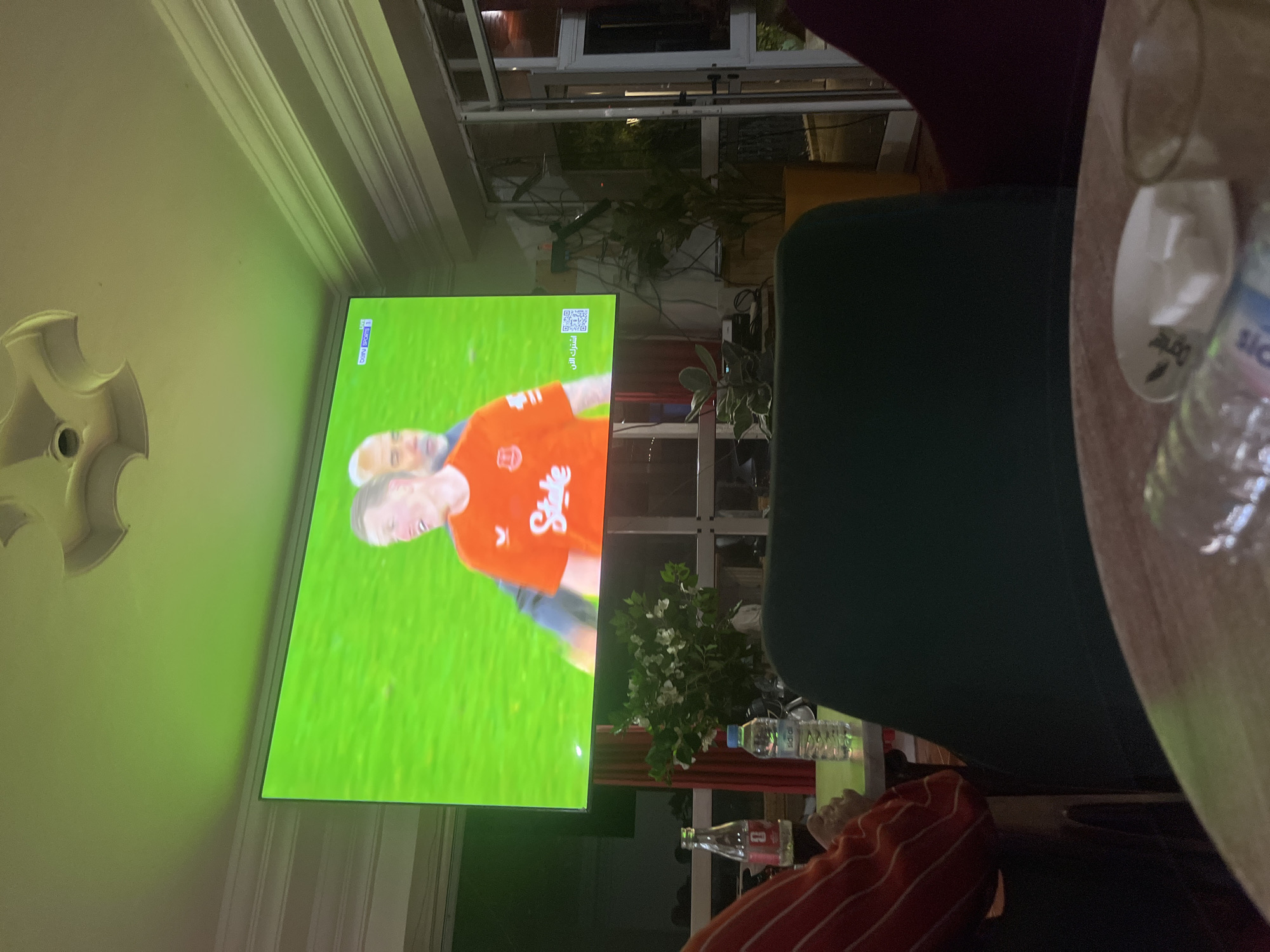 The scene shows a television screen displaying a football (soccer) match. A person is visible on the screen wearing an orange jersey. The TV is mounted on a wall with decorative molding in what appears to be a living area. A green upholstered chair is visible in the foreground. A table with a drinking glass and bottle is also present. Through a window or glass enclosure, greenery is visible. The location is Ouaouizert, Morocco.