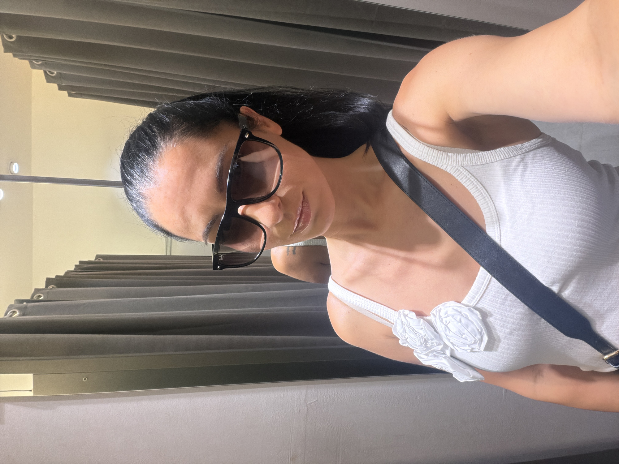 A woman with dark hair pulled back and wearing sunglasses and a white tank top with floral embellishments on the strap stands in what appears to be a fitting room. The background consists of gray curtains and a mirror reflecting her left shoulder and arm. The setting is indoors, and there are no immediate indications of the city of Pleven, Bulgaria.