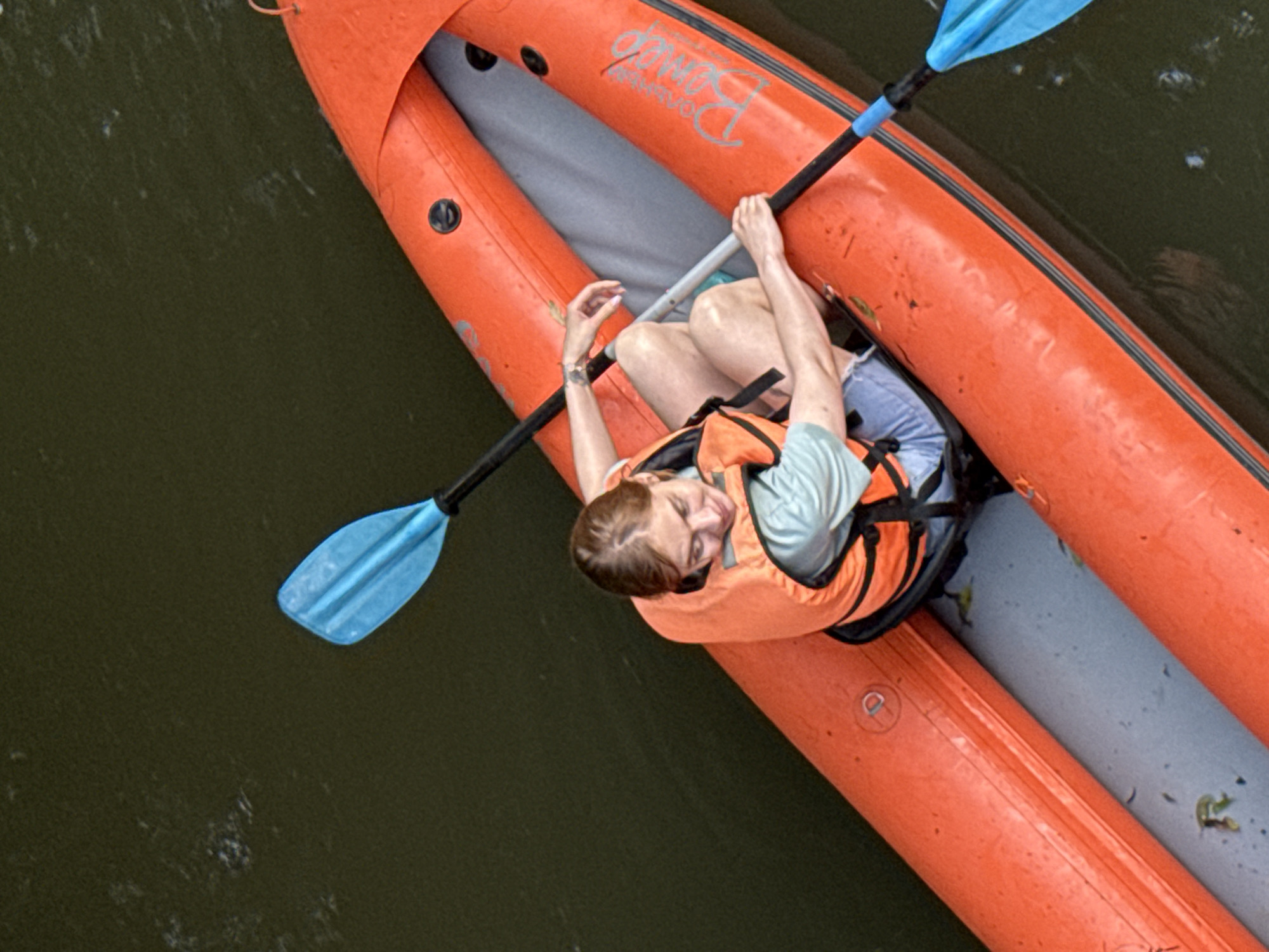 The image shows a person in an orange inflatable kayak on a body of water. The person is wearing a light blue t-shirt and an orange life vest. They are holding a blue and black kayak paddle. The kayak has the text "BOMBO" visible on its side. The water is dark green and appears somewhat murky. The scene suggests a recreational activity like kayaking, likely taking place during daylight hours given the lighting. The person's head is tilted back and their eyes are closed, which could indicate relaxation or enjoyment. The specific location cues for Moscow are not present in the visual information.