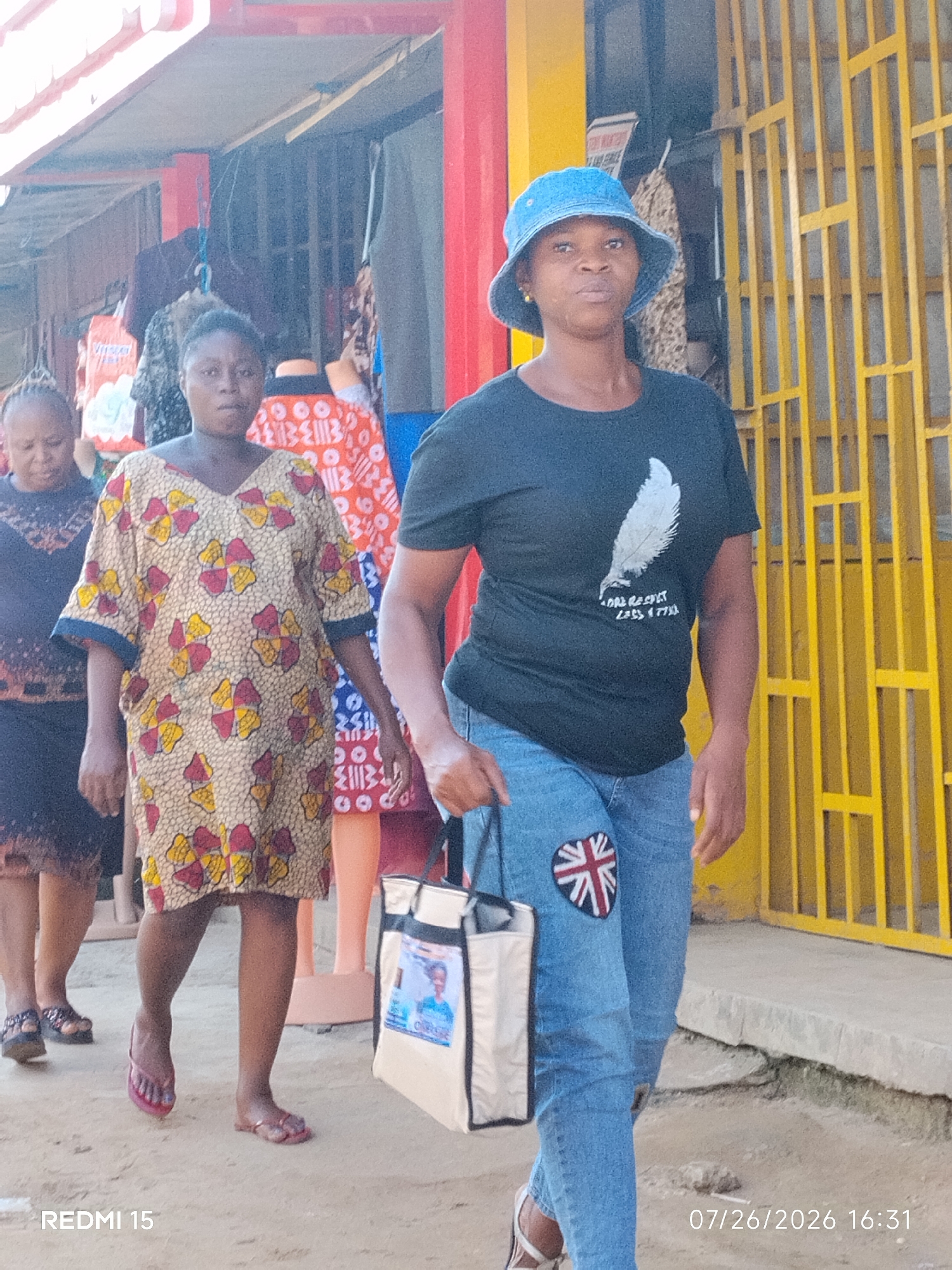 In Umuode, Nigeria, three women walk along a sandy street lined with shops. The woman in the foreground wears a blue bucket hat, a black t-shirt with a white feather graphic, and blue jeans adorned with a Union Jack patch on the left thigh. She carries a light-colored tote bag featuring a picture of a young boy in a suit. To her left, a woman in a vibrant, patterned yellow and red dress walks with her head bowed. Behind her, another woman in a dark dress is partially visible. The street features a yellow metal grate on the right and various clothing displays visible in the shopfronts. The image is dated 07/26/2026 16:31.