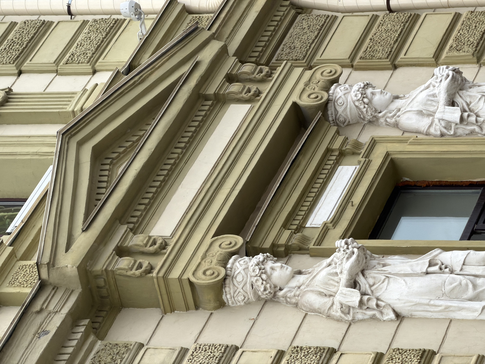 This is a close-up, low-angle shot of an ornate building facade in Nizhny Novgorod, Russia. The primary subjects are two white relief sculptures of women, each lying on their side and adorned with intricate headwear and flowing garments. Their faces are depicted in profile, with their eyes closed and expressions serene, suggesting a state of repose or contemplation. The sculptures are integrated into the architectural design of the building, which features a combination of light green and cream-colored elements.
The architectural details are elaborate, including decorative carved panels with textured inserts, fluted pilasters, and geometric moldings. The sculptures are positioned below windows, one of which is visible in the upper right portion of the image, reflecting a bright, possibly overcast sky. The overall aesthetic points to a historical or neoclassical architectural style. The lighting suggests it is daytime, but the specific time of day is not discernible. There is no visible text in the image. The scene captures the artistic and architectural embellishments of an older building, highlighting the craftsmanship of the sculptures and the surrounding facade.