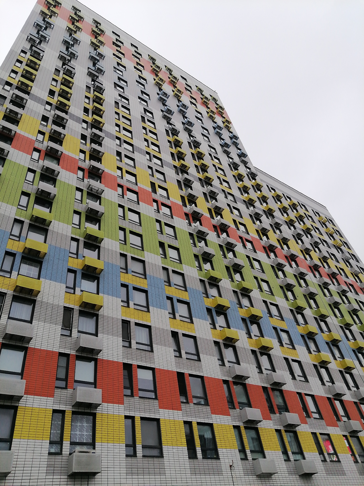 A low-angle shot depicts a tall, modern apartment building in Moscow, Russia. The building's facade is characterized by horizontal bands of colorful tiles in hues of orange, yellow, green, and blue, interspersed with grey-and-white sections. Windows with black frames punctuate the facade, and a gray box-like projection, presumably an air conditioning unit or a balcony extension, is situated beneath most of the windows. The sky visible in the upper portion of the frame is overcast and grey.