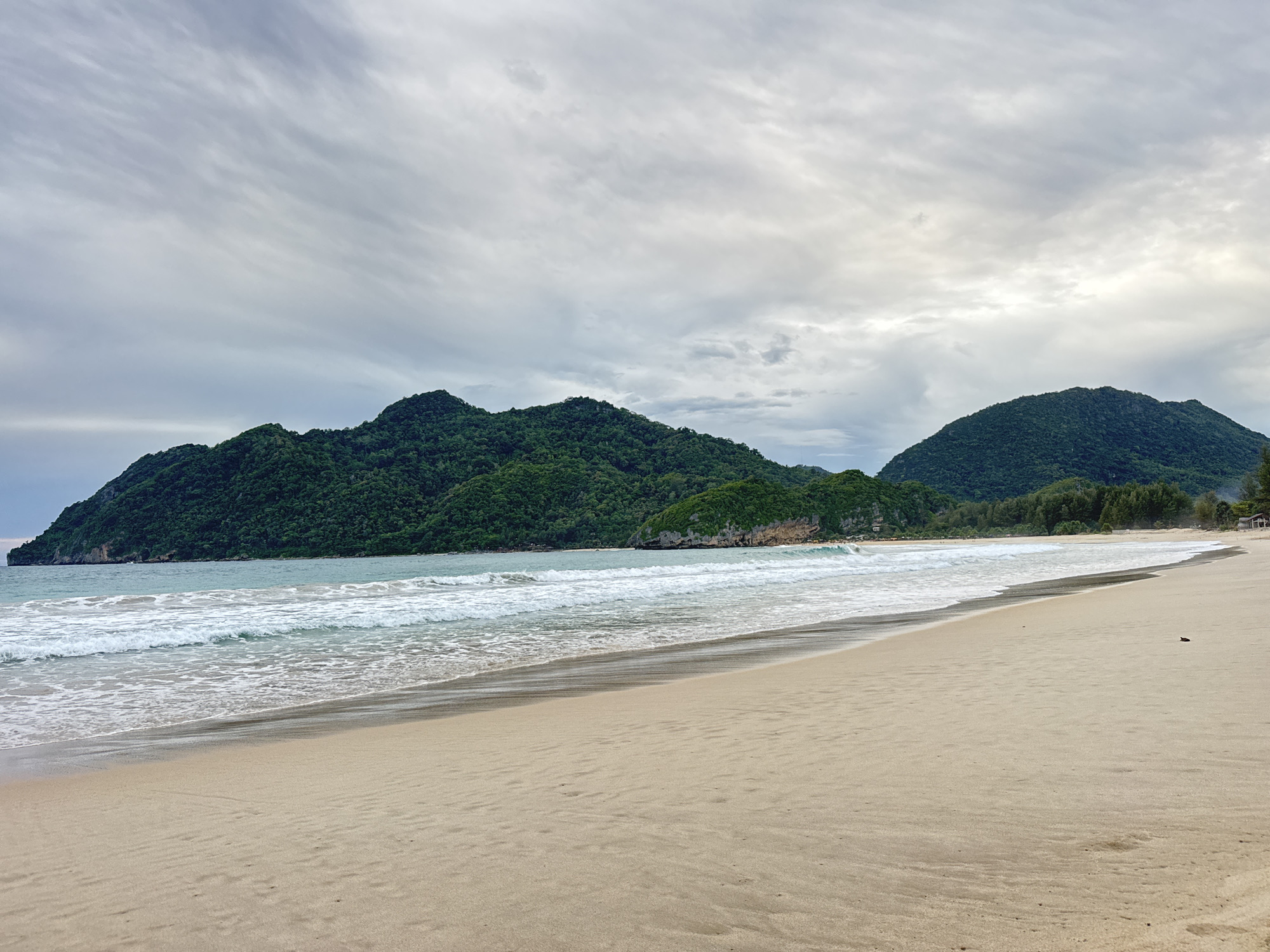 The image depicts a serene beach scene in Meunasahbale, Indonesia. The foreground is dominated by a wide expanse of light brown sand, showing subtle ripples and a few small indentations. A dark, wet line marks the transition from the dry sand to the damp area closer to the water.
The middle ground features the gentle turquoise ocean with white-capped waves rolling towards the shore. The water appears calm, with a shallow depth near the beach.
In the background, lush green, jungle-covered mountains rise dramatically from the coastline, creating a striking contrast with the soft sand and blue water. The sky is overcast with a gradient of grey and white clouds, suggesting a possibly humid or pre-rainy atmosphere. The overall ambiance is peaceful and natural, with no visible people or man-made structures directly in the foreground or mid-ground, though a hint of buildings or structures might be present in the far distance along the coastline.