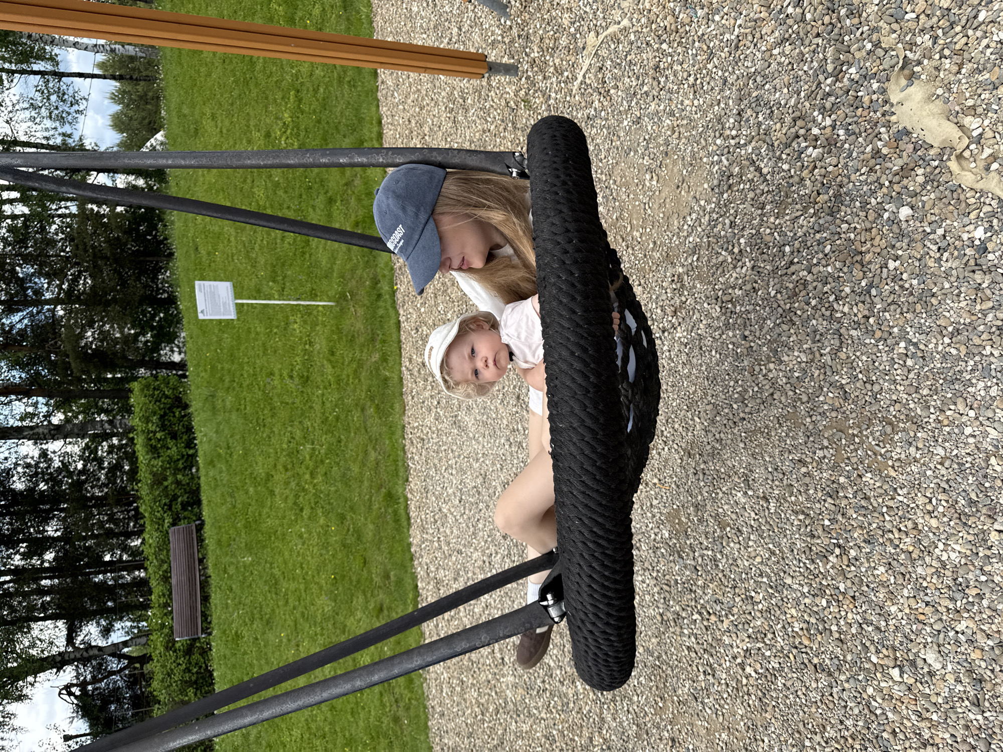 The image displays a peaceful outdoor scene, likely a park or playground in Dobrograd, Russia, during clear daytime. A young woman with long blonde hair, wearing a white t-shirt, light-colored shorts, and a blue baseball cap (with the word "COAST" visible), is seated in a large black tire swing. She holds a baby or toddler on her lap, who wears a light-colored top and a white sun hat.
The woman gazes tenderly at the baby, while the baby looks towards the viewer with a calm expression. The swing is supported by black metal poles, and the tire is wrapped in black rope. The setting features a neatly mown green lawn on the left and a gravel path on the right. In the background, trees line the edge of the grass, and a brown wooden bench is visible. A small white informational sign on a pole stands on the lawn, but its text is unreadable. The ground is covered in light-colored pebbles and gravel. The scene conveys a quiet moment of leisure and familial interaction in a pleasant outdoor environment.
