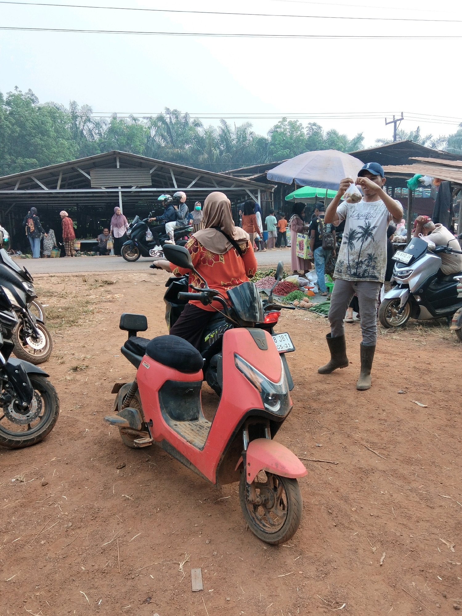 A street market scene in Tirto Sari, Indonesia, features several people and parked motorcycles on a dirt ground. In the foreground, a red electric scooter is parked facing left, with its front wheel and fender visible. To the left, the front wheel and part of the body of a black motorcycle are visible. In the mid-ground, a woman wearing a headscarf and an orange patterned shirt rides a black motorcycle. To her right, a man wearing a white t-shirt with palm tree graphics, grey pants, and dark boots is holding up a clear plastic bag with food items. He is facing the camera. Behind him, a silver motorcycle is parked. In the background, several stalls with open roofs are visible, with people browsing and shopping. A large umbrella provides shade over some of the stalls. The environment appears to be an outdoor market with a backdrop of trees and a bright, overcast sky.