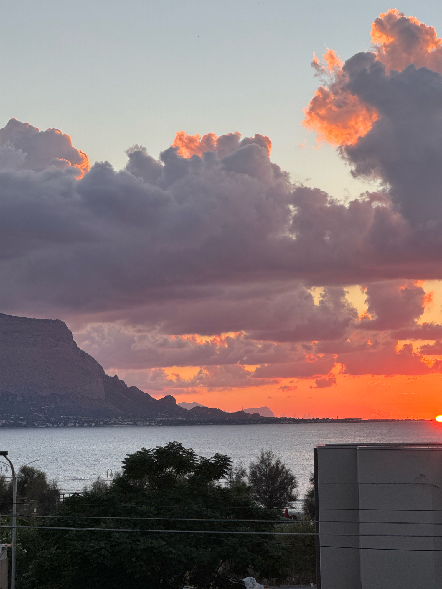 The image depicts a sunset over the sea, with dramatic clouds painted in shades of orange, pink, and gray. A large, mountainous coastline stretches across the left side of the frame, with a town nestled at its base and a body of water in the foreground. A modern building occupies the lower right portion of the image, its stark gray facade contrasting with the natural beauty of the sunset. Dark green trees and power lines are visible in the mid-ground, adding a touch of urban interference to the otherwise serene scene. The sun, a fiery orb, is setting on the right horizon, casting a warm glow across the water and sky. This view is characteristic of the coastline near Capaci, Italy, a town known for its picturesque scenery. There are no people or animals visible in the image.