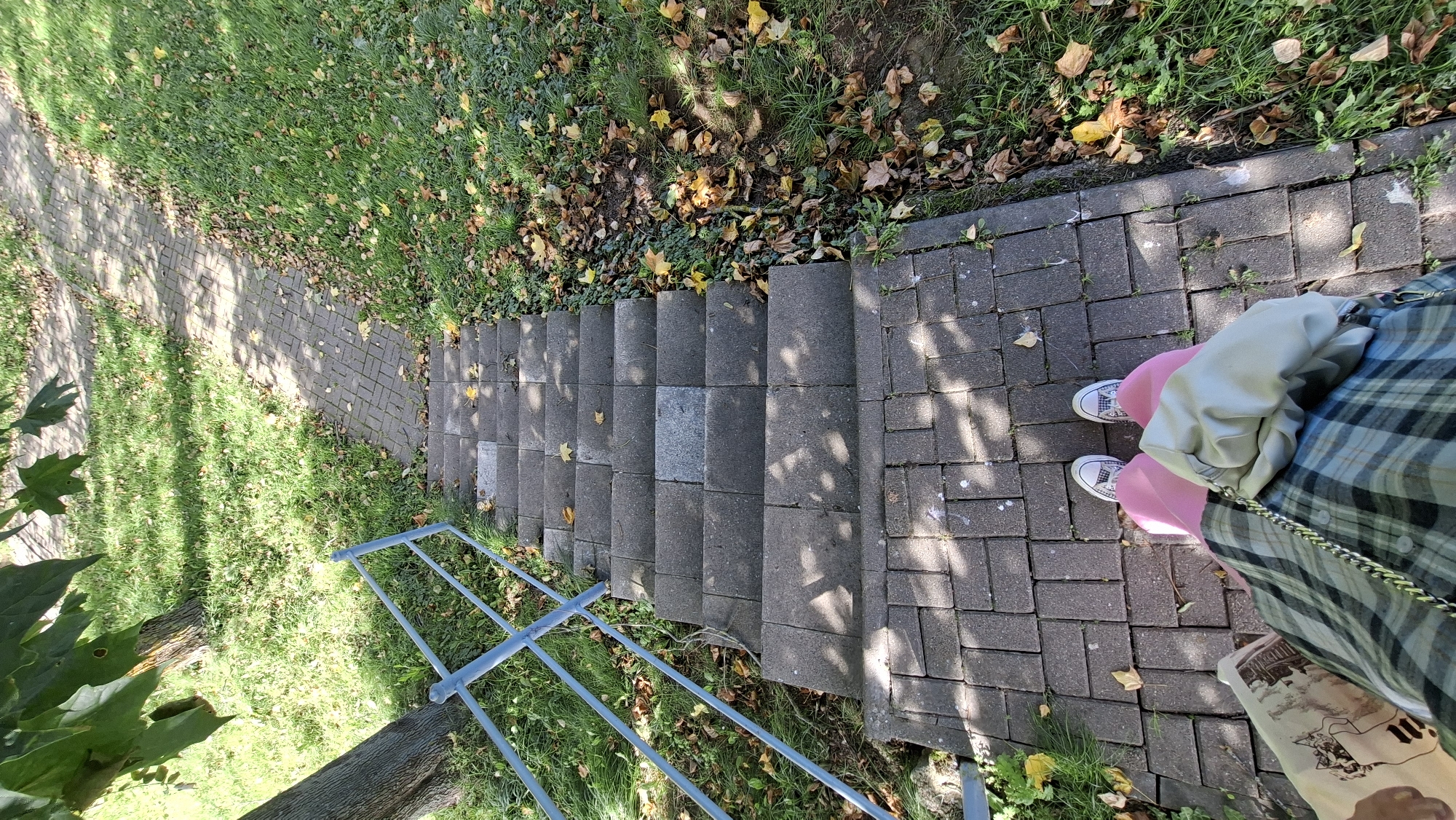 An overhead perspective captures a person standing on a brick pathway at the bottom of a set of concrete stairs. The individual is wearing white Converse sneakers and pink pants, with a plaid skirt and a light-colored purse draped over their arm. The pathway and stairs are bordered by green grass interspersed with fallen autumn leaves. A light blue metal handrail runs along the left side of the stairs. The scene is illuminated by sunlight, creating shadows from trees onto the pathway and stairs. The location is identified as Myadzyel, Belarus.