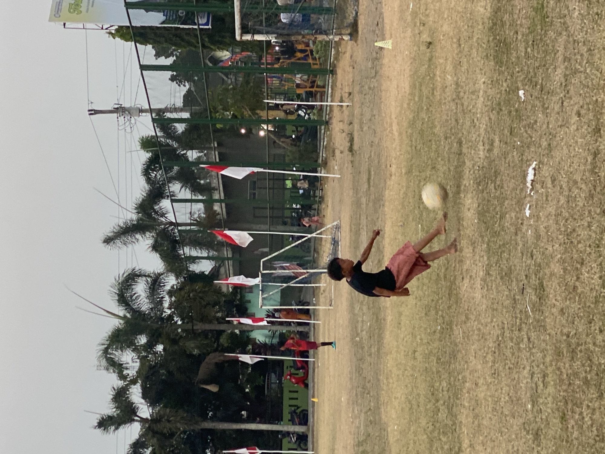 A young boy is captured in mid-kick, striking a soccer ball on a dry, grassy field. He is wearing a black t-shirt and reddish-brown shorts. His body is angled towards the left of the frame, with his right leg extended to connect with the ball, which is in motion. In the background, several Indonesian flags are planted on poles. Behind the flags, a fence encloses a sports complex area, with a soccer goal visible on the right. Palm trees and other vegetation line the perimeter of the field, with buildings and a utility pole visible in the distance. The sky is overcast. The scene appears to be taking place in the city of Kasihan, Indonesia.