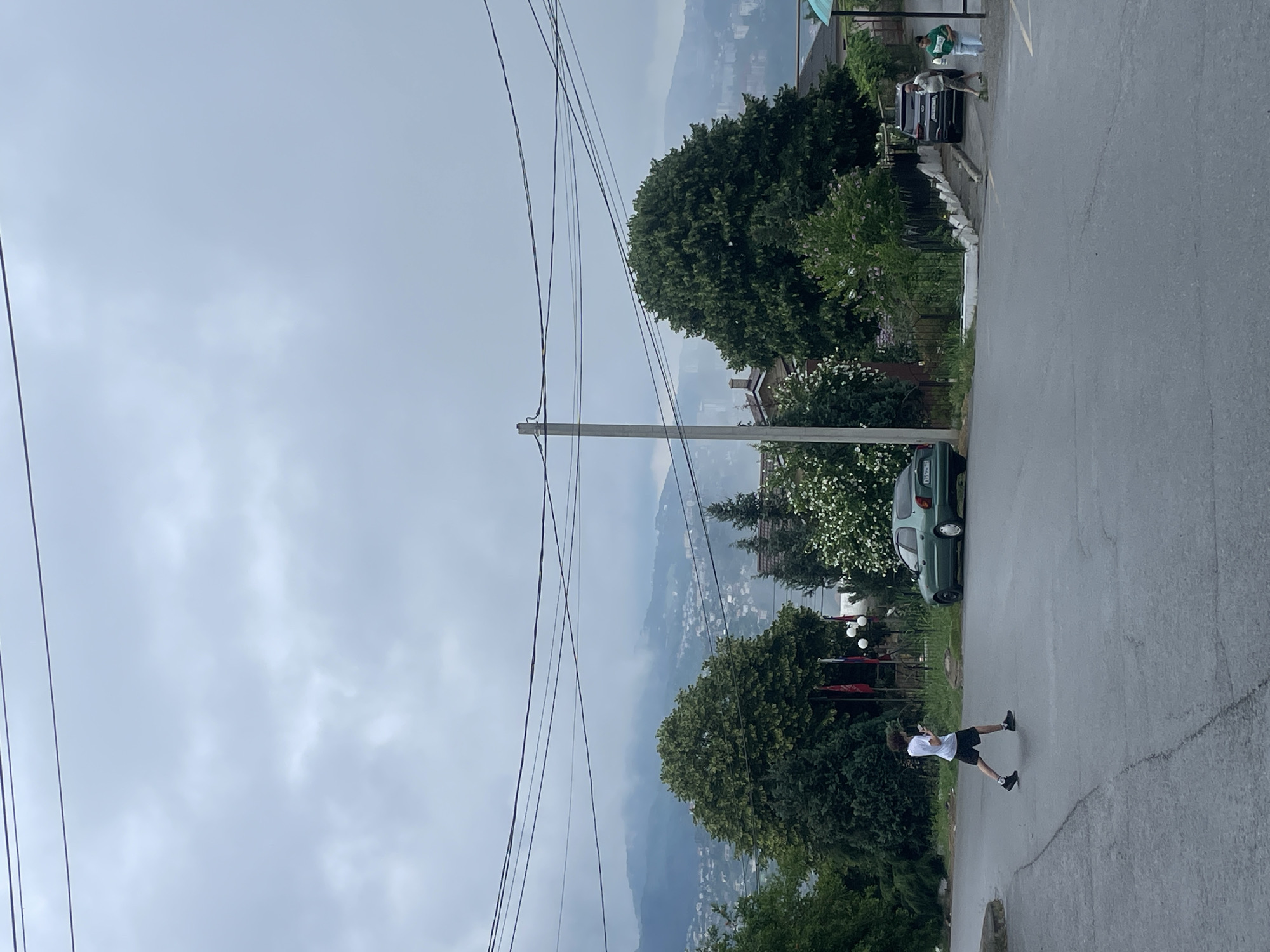 A person is walking across a wet road in a suburban area, possibly Baranovka, Russia, under an overcast sky. The person, wearing a white t-shirt and black shorts, is looking down at a mobile device. To the left of the person, a green sedan is parked on the side of the road. In the background, trees and houses are visible, with a hazy mountain range in the distance. Several power lines are strung across the sky. Further down the road, two individuals, one in a light-colored shirt and shorts and another in a green t-shirt and blue pants, are walking towards the right side of the frame. A dark SUV is parked near them. The environment appears to be a typical residential street with elements of nature and urban infrastructure.