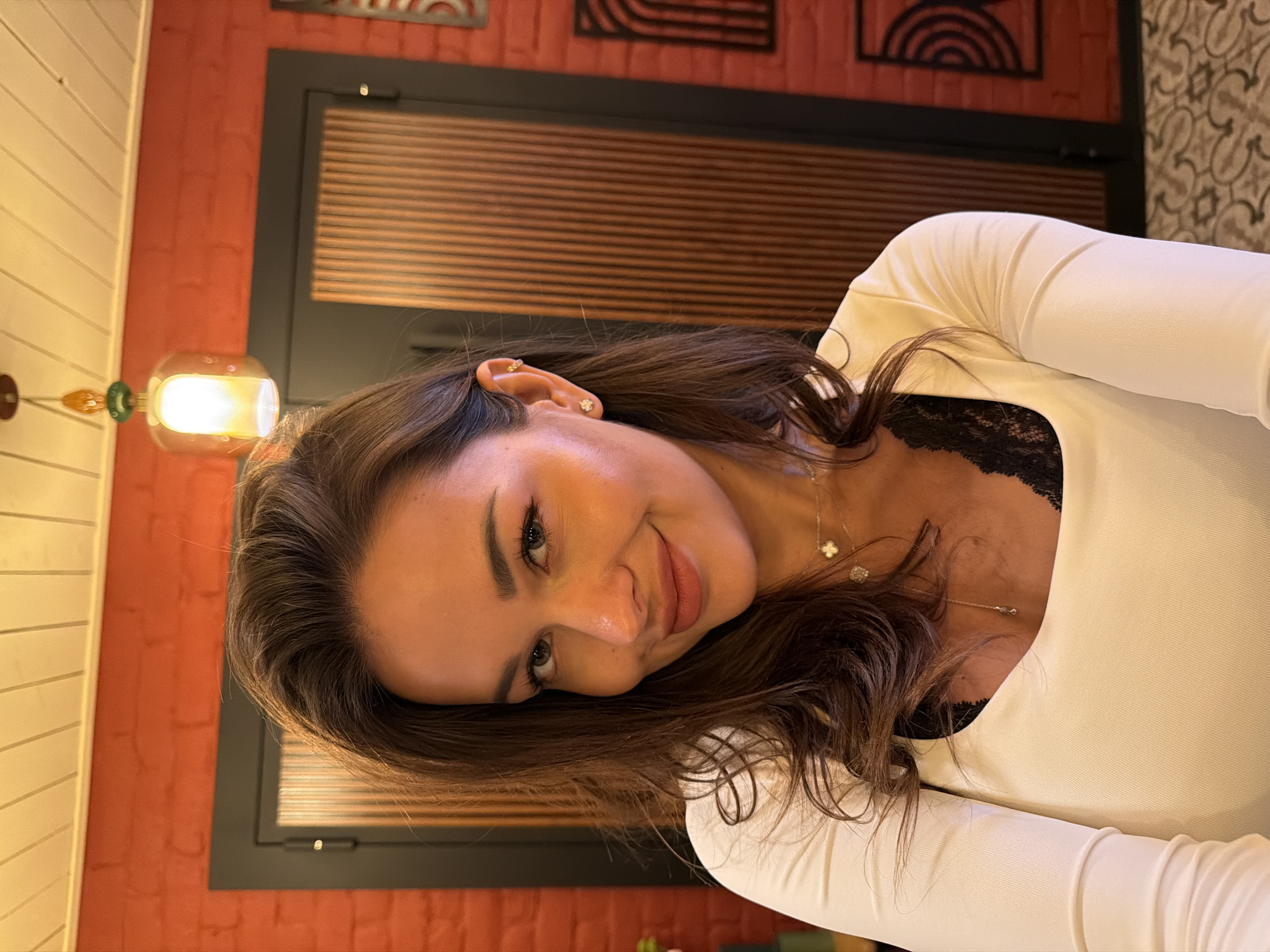 A medium shot photo shows a woman with long, dark, wavy hair and light skin. She is smiling and looking towards the camera. She is wearing a white long-sleeved shirt with a black lace trim visible at the neckline, and a necklace with a clover pendant. She has small earrings in her ears. The background consists of a red brick wall, a wooden door, and a light fixture. The scene appears to be indoors, possibly in a restaurant or cafe. There are no discernible elements that would indicate the location as Saint Petersburg, Russia.