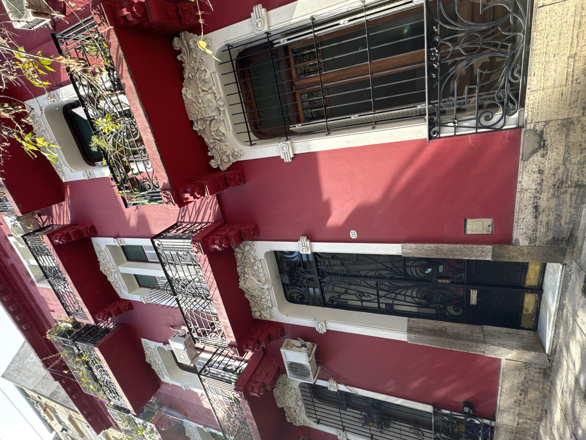The image shows a red building in Buenos Aires, Argentina, with white trim around windows and doors. The building has multiple stories and each floor has wrought iron balconies. The windows on the upper floors have decorative metal bars. An air conditioning unit is attached to the wall below a window on one of the lower floors. The building has a main entrance door with ornate ironwork. The building number is visible near the door. There are trees and sunlight visible in the background, suggesting the image was taken during the day.