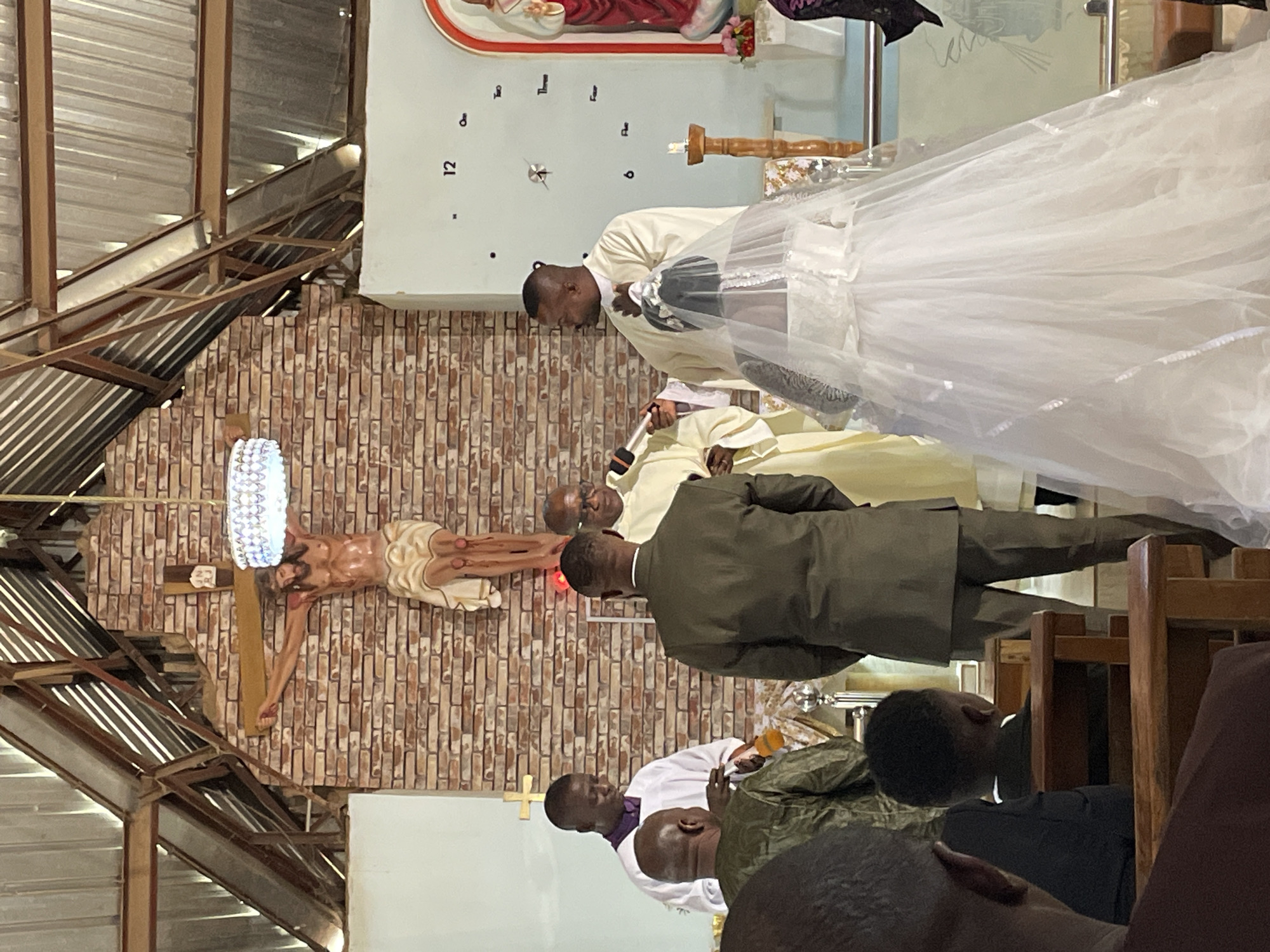 A wedding ceremony is taking place inside a church with exposed metal beams. A crucifix hangs on a brick wall near the altar. A bride in a white wedding dress and veil stands beside a man in a white robe and collar holding a microphone. Another man in a green suit stands between them and a man in a light robe. Pews filled with people sit in the foreground. A clock-like sculpture hangs in the background. It is not possible to determine the specific location in Jos, Nigeria from this image.