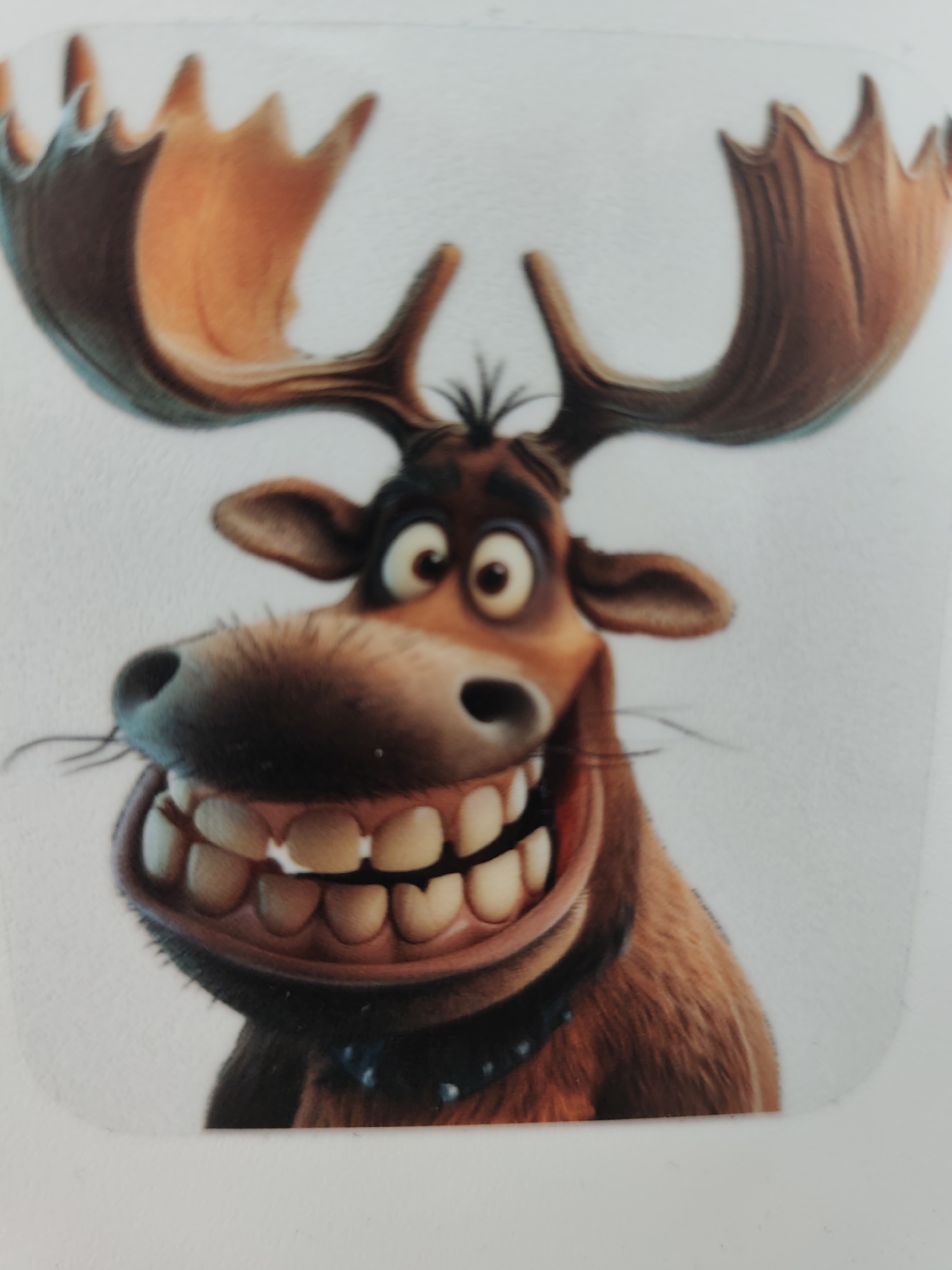 The image presents a head-and-shoulders portrait of a brown anthropomorphic moose character, identified as being in the city of Pazardzhik, Bulgaria. The character is rendered in a bright, animated style against a plain white background. It features large, palmate antlers with variations of brown and orange hues, curving broadly upwards. The moose's face displays wide, round eyes with dark pupils, a broad dark snout, and fine dark whiskers. Its mouth is open in an exaggerated, wide grin, prominently showcasing two full rows of large, bright white, uniformly shaped teeth. The fur on its body and neck appears brown with a soft, textured appearance, and a dark, studded collar is visible around its neck.