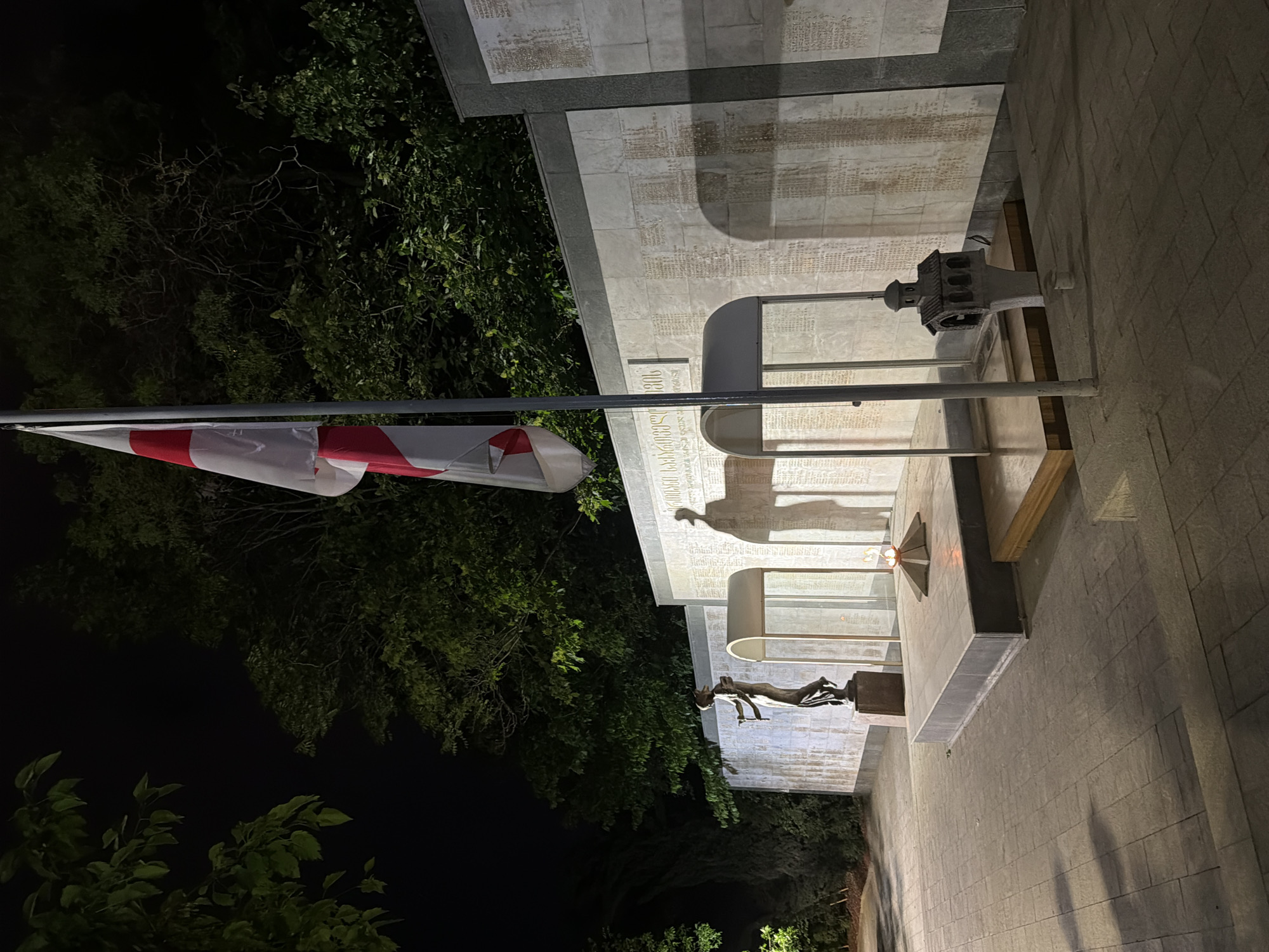 The image depicts a memorial in Tbilisi, Georgia, at night. The monument comprises a stone structure with inscribed plaques and a small sculpture of a man on a pedestal. A Georgian flag is visible on a pole extending from the structure, juxtaposed against the dark backdrop of trees and sky. The area surrounding the monument is paved with stone bricks, suggesting a public space.
