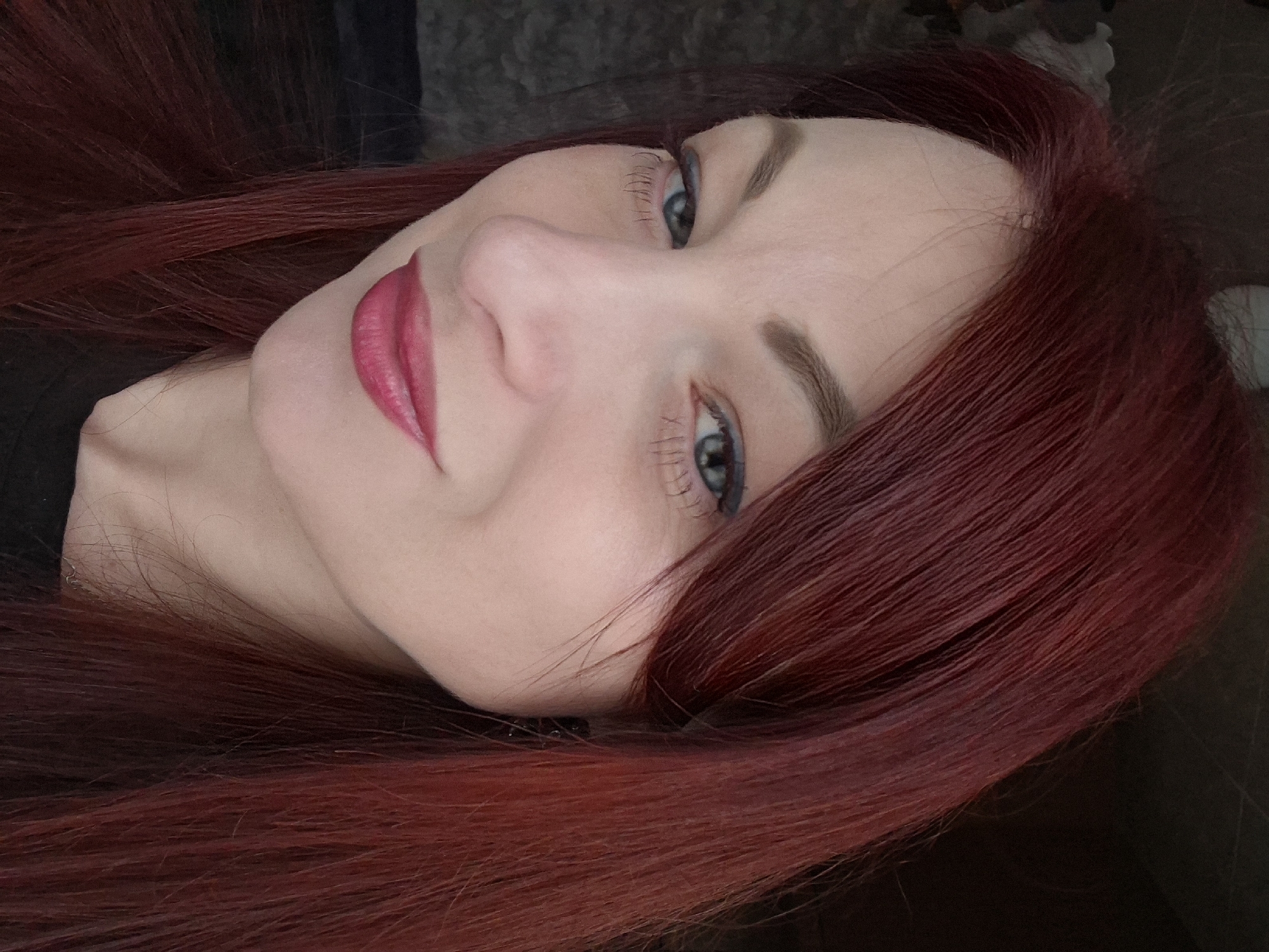 A close-up, angled shot of a person with fair skin and red hair. The person is facing the camera, with their head tilted to the right. They have blue eyes and are wearing red lipstick. The background is dark and blurry. The location is Unknown, Unknown.