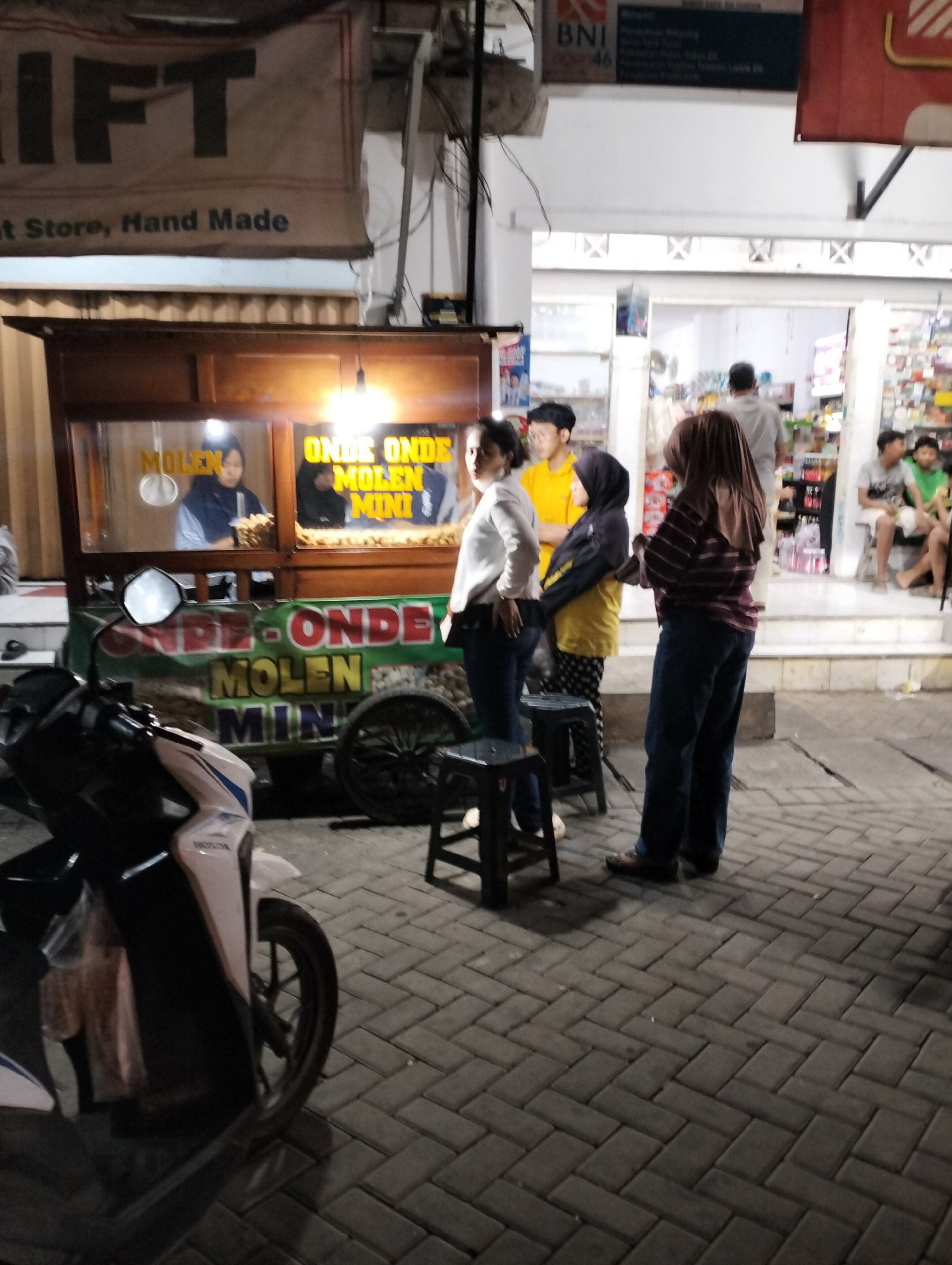 The image depicts an outdoor scene at night in Semarang, Indonesia, likely a street or market area. In the foreground, a white and blue motorcycle is parked on a herringbone-patterned brick pavement. Behind the motorcycle is a food cart selling "Onde Onde Molen Mini." The cart has a wooden frame and glass display windows. Inside the cart, two individuals are visible, one wearing a blue headscarf and the other a dark headscarf. They appear to be preparing or serving food.
To the right of the food cart, a group of people are standing. One person is wearing a yellow shirt and another a dark hijab and jacket. A woman in a white long-sleeved shirt and blue jeans is standing to the left, facing away from the camera. Further in the background, there is a building with shelves stocked with items, suggesting it might be a small shop or minimarket. Several individuals are visible inside and outside this establishment.
Above the food cart, a large banner with the word "RIFT" is prominently displayed, along with smaller text that reads "t Store, Hand Made." There are also signs with Indonesian text and a logo for BNI (Bank Negara Indonesia). The lighting appears to be artificial, with streetlights and lights from the food cart and the shop illuminating the scene. The overall atmosphere suggests a bustling commercial area at night.