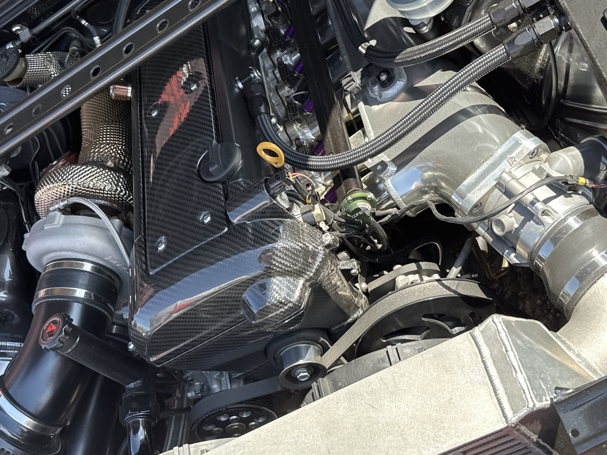 The image shows a high-performance car engine bay. Prominent features include a carbon fiber valve cover, a turbocharger with heat wrap, a billet aluminum intake manifold, fuel injectors with purple anodizing, and various braided stainless steel hoses and lines. The engine is belt-driven, with visible pulleys and a serpentine belt. An intercooler is visible at the bottom of the image. There is no indication of location or presence of any people.
