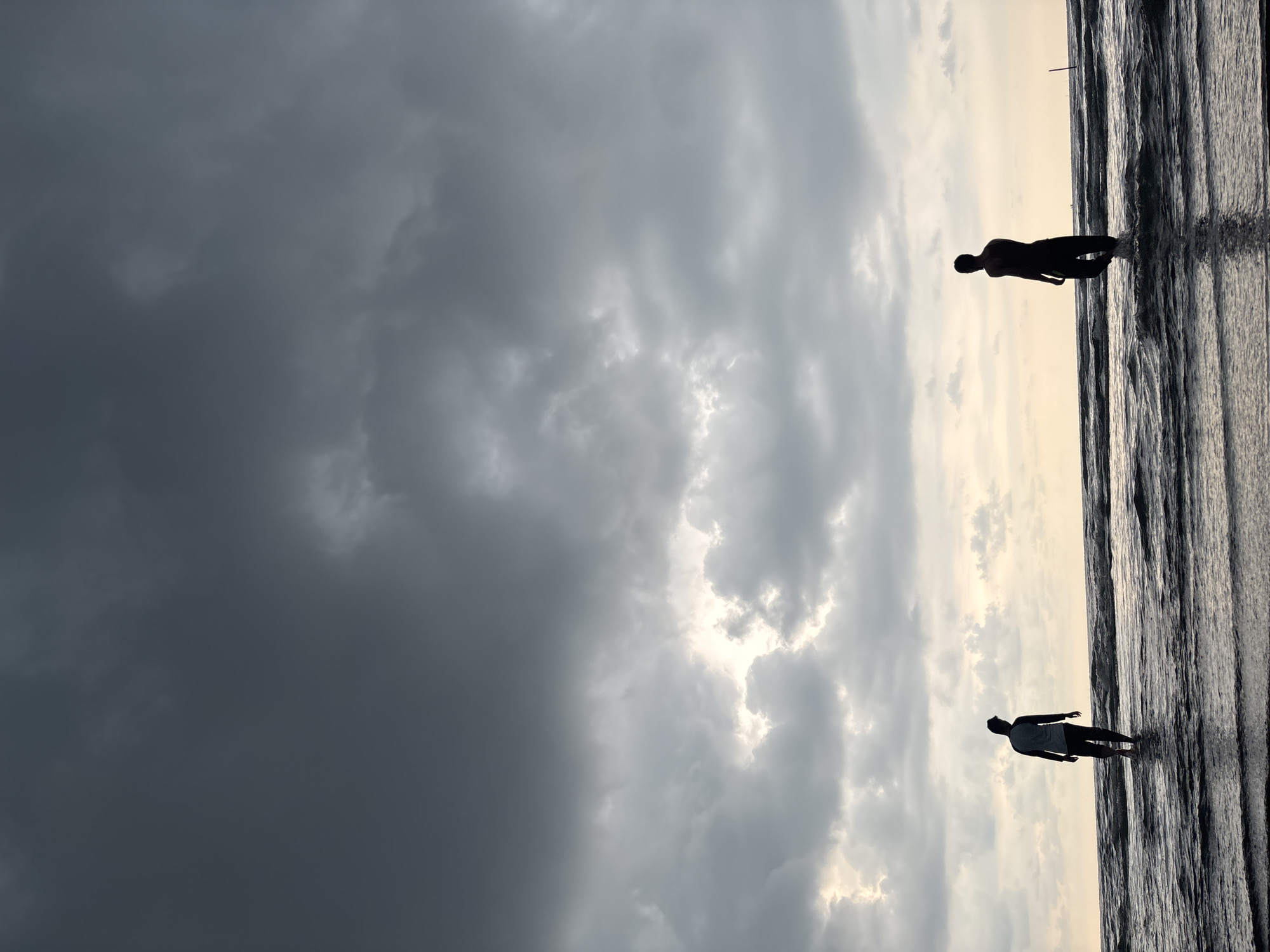 The image captures a coastal scene, likely a beach, under a dramatic sky filled with storm clouds. Two silhouetted figures are visible along the shoreline. One figure stands in the water near the top edge of the frame, while the second is positioned lower down. The environment consists of a dark, turbulent sea meeting a sky characterized by varying cloud formations. The location appears to be a beach in Angkleng Barat, Indonesia.