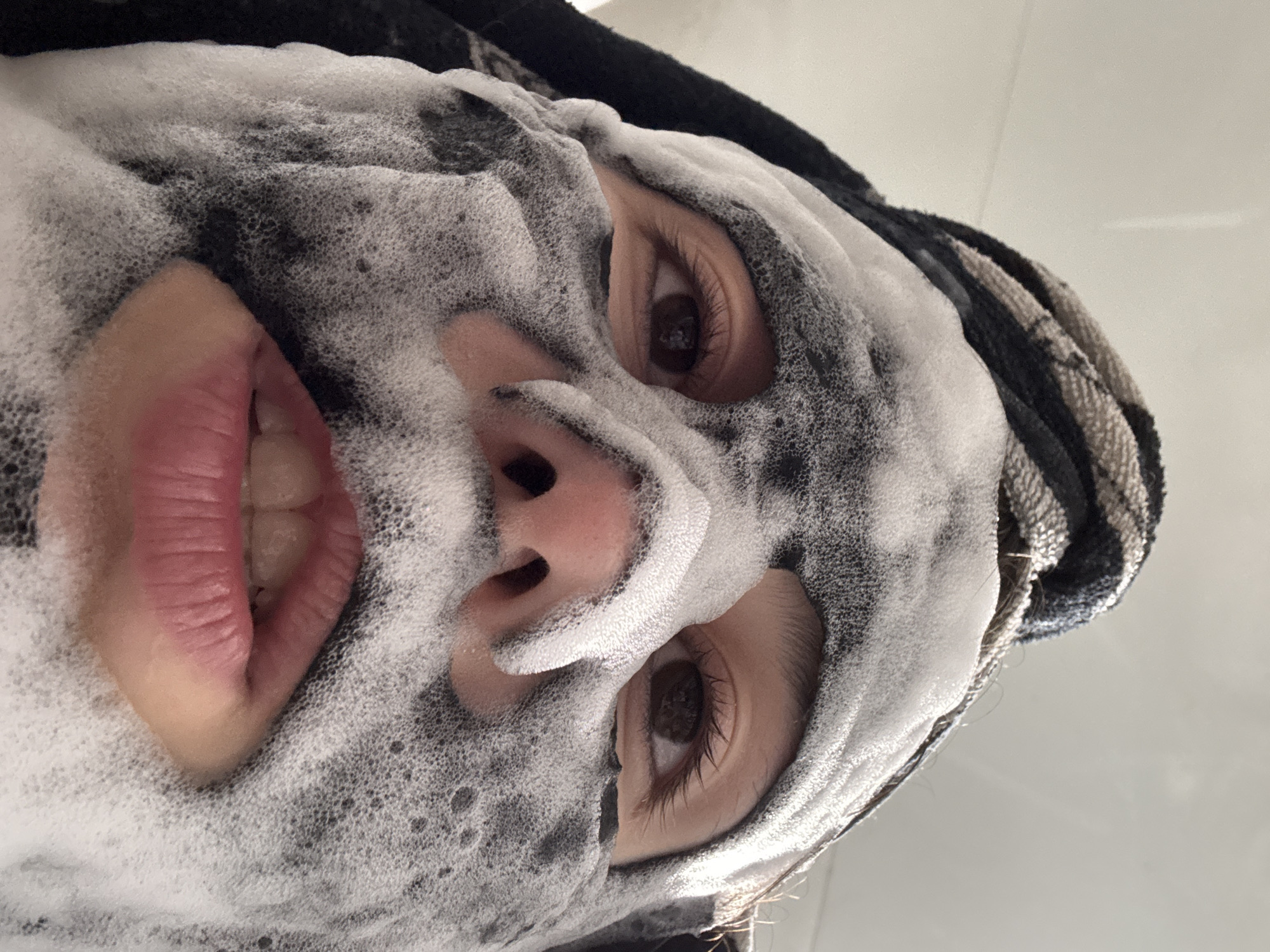 A close-up, tilted upward selfie shows a person with a facial cleanser that is black and foamy with white bubbles covering most of their face. The person has brown eyes and is slightly smiling, revealing their upper teeth. A striped black and white towel or headband is visible at the top of their head. The background is a plain, light-colored wall or surface. There is no indication of the location being in Фоки, Russia.