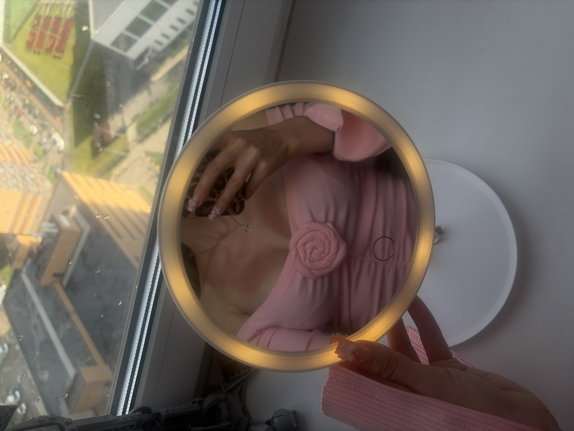 In the foreground of the image, a person's hand, adorned with pink nail art featuring small white bows and white polka dots, is holding a piece of pink textured fabric. This hand is positioned to the left of a round, illuminated mirror. The mirror reflects the upper body of a person wearing a pink off-the-shoulder top with a rose-like embellishment in the center and a delicate silver cross necklace. The background visible through a window shows a cityscape with multiple buildings, some appearing residential and others industrial, under a bright sky. The windowpane has water droplets on it, suggesting recent rain or humidity. The overall scene is indoors, on a windowsill, overlooking an urban environment.