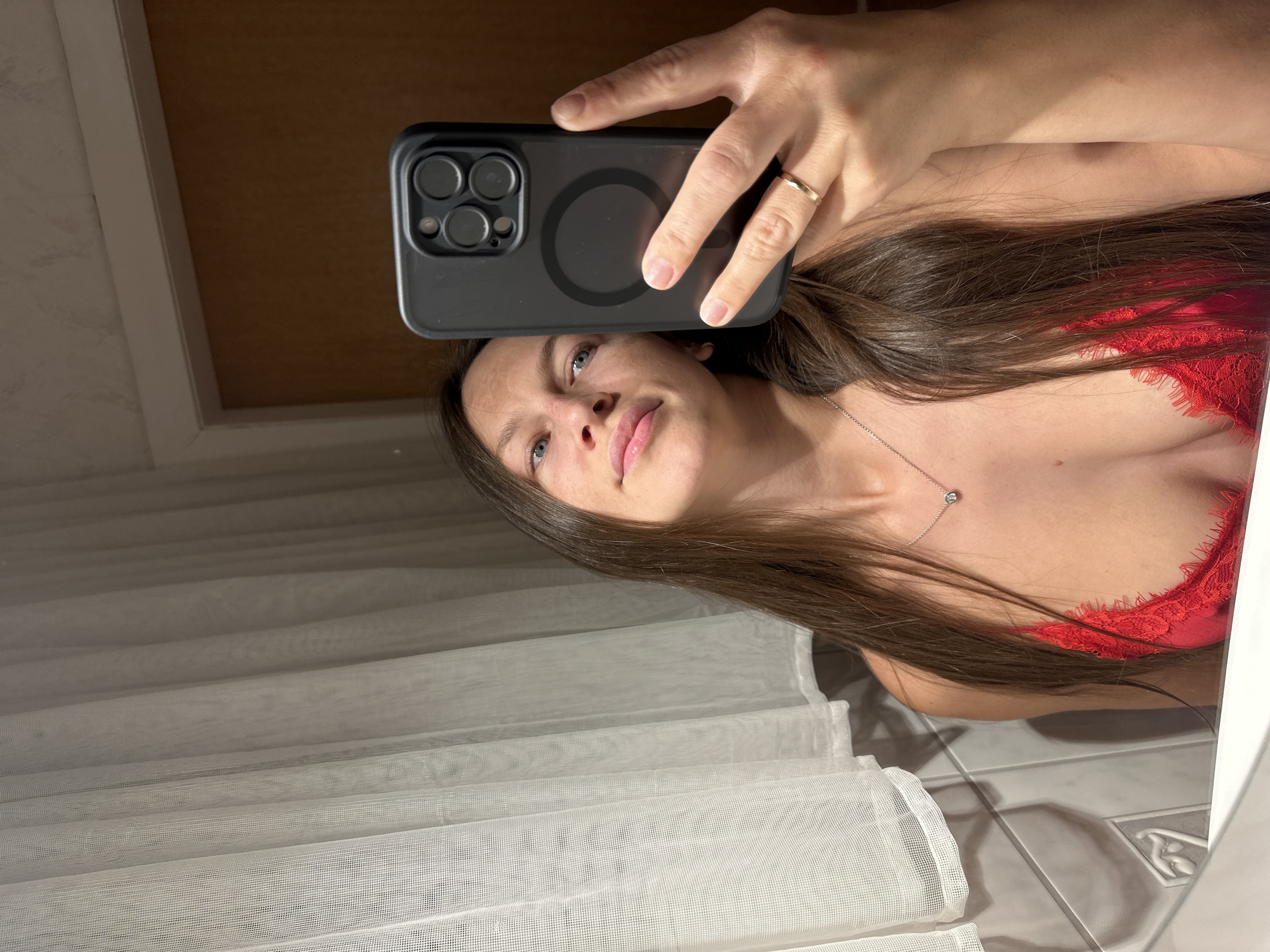 An adult female takes a mirror selfie in an indoor setting, identified as Fredenbeck, Germany. She has long, straight, dark brown hair, blue eyes, and fair skin with minor freckling on her nose and forehead. She is wearing a red, lacy-edged, low-cut top, a silver chain necklace with a small clear stone pendant, and a simple gold band ring on the ring finger of her right hand. Her right hand holds a black smartphone, featuring a multi-lens camera array and a circular MagSafe imprint on its back. The background, reflected in the mirror, displays a sheer white pleated curtain on the left, below which are white rectangular wall tiles with subtle decorative relief. To the right, a light brown wooden door or wall panel is visible, topped by white molding and a light-colored textured wallpaper. The bottom edge of the mirror frame is also visible, appearing white.