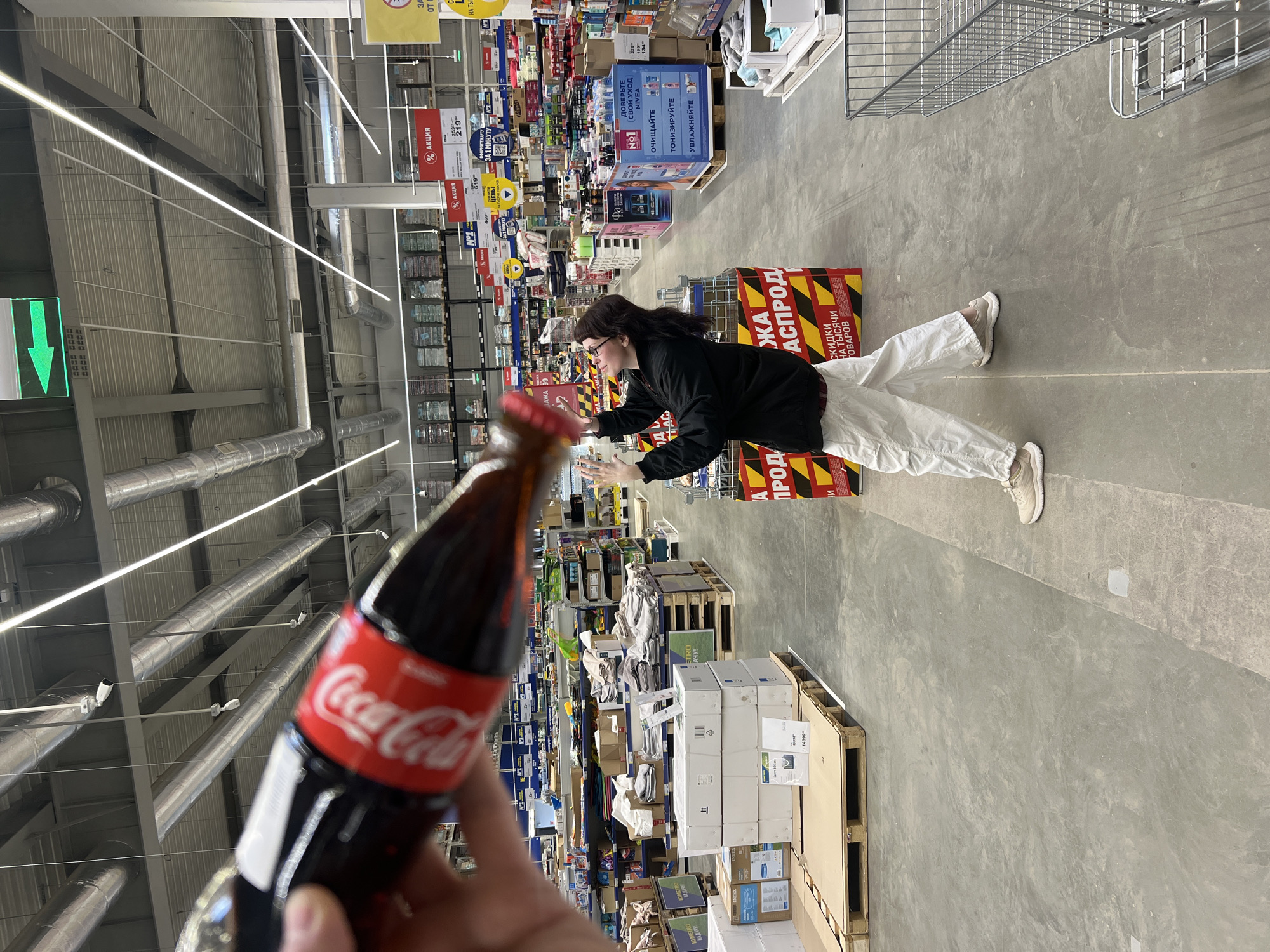The photo shows a Coca-Cola bottle being held in the foreground. In the background, a woman is lying on a stack of pallets in the aisle of a large retail store. The aisles are stocked with a variety of goods. Signs above the shelves indicate sales and prices. The location is likely a store in Izhevsk, Russia, based on Cyrillic text on signs and products. The woman appears to be posing.