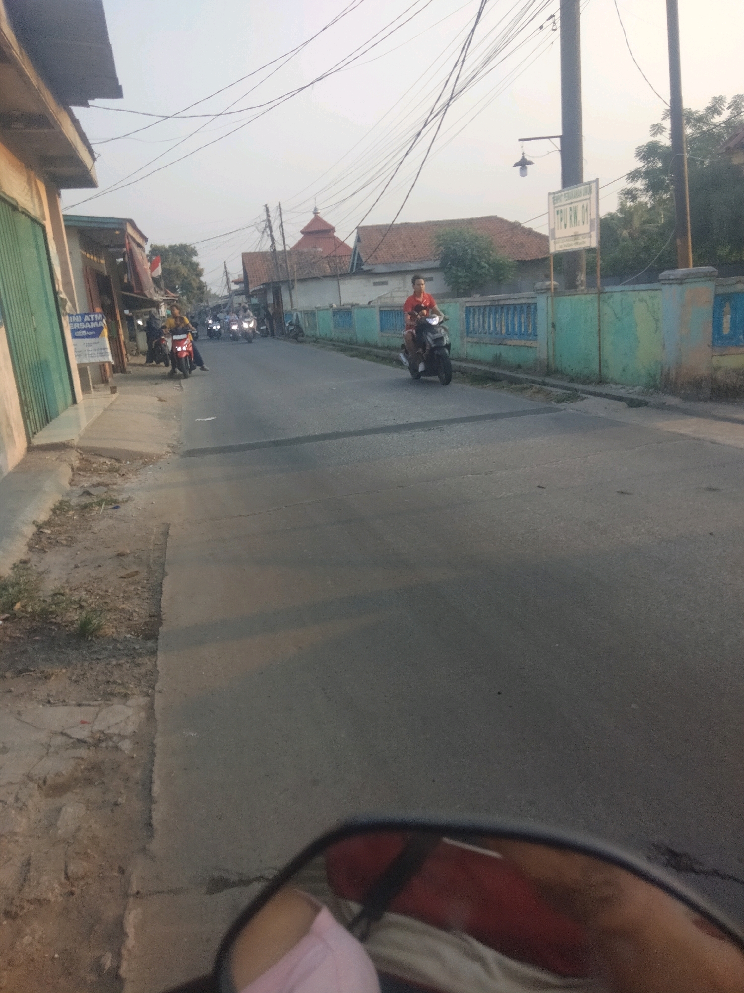 This is a horizontal, eye-level, medium shot of a street in Pangadegan, Indonesia, likely taken from the perspective of someone riding a motorcycle. The sun is low, casting long shadows and bathing the scene in a warm, golden light. The road is asphalt and appears to be two lanes wide, with a paved sidewalk on the left. Several motorcycles are visible on the road, some with riders.
On the left side of the road, there are buildings with shops. One shop has a green metal gate and a sign that reads "INI ATM BERSAMA". Further down, the buildings give way to a more open area with trees.
The right side of the road features a low, turquoise-colored wall with blue trim. Beyond the wall, there are more trees and buildings. A prominent feature on the right is a pole with a street light and a white sign that reads "TPU RW. 01". A red-roofed mosque with a dome is visible in the distance, behind the buildings and trees.
A man in a red shirt is riding a dark-colored motorcycle in the middle of the road, heading towards the viewer. Other motorcycles are further down the road, creating a sense of movement and activity. The sky is pale and overcast, with several power lines crisscrossing it.
In the foreground, a motorcycle's rearview mirror is visible, reflecting a portion of the rider's face and shoulder, as well as a pink garment. The overall impression is of a typical street scene in a residential or semi-commercial area of an Indonesian town during late afternoon or early morning.