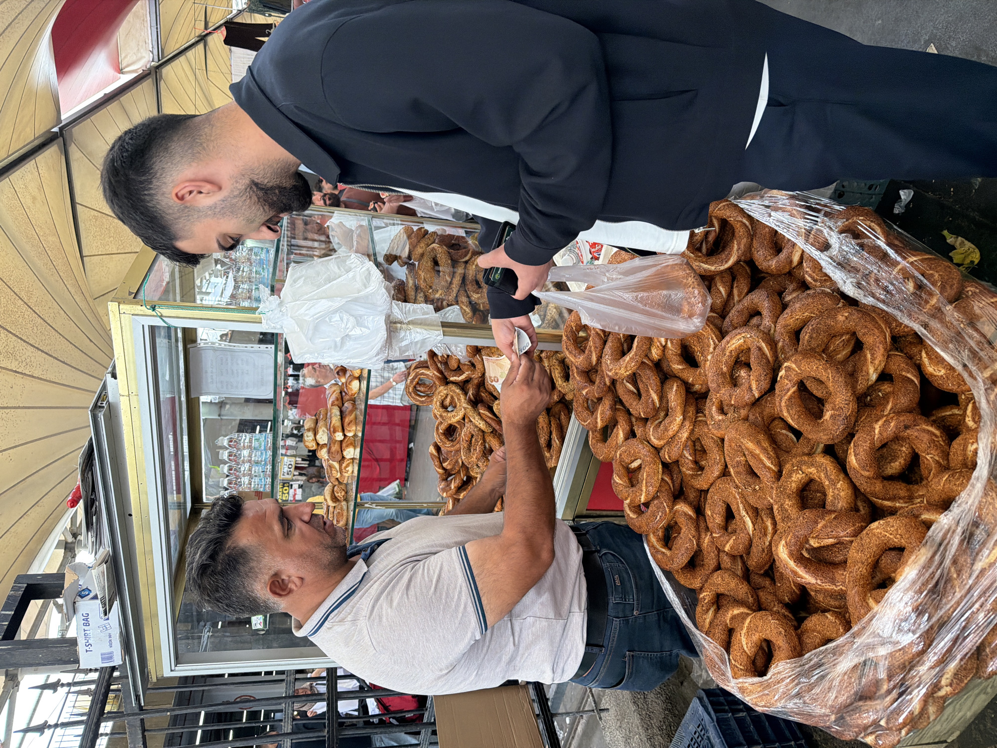A man in a gray polo shirt and jeans is at a street food stall in Kadıköy, Turkey, selling simit, a type of Turkish bagel. He is handing a simit to a customer, a man in a black jacket and dark pants, who is paying with cash. The stall is piled high with simit, and there are bottles of water and other food items displayed behind the glass. The scene appears to be a busy market or shopping street.