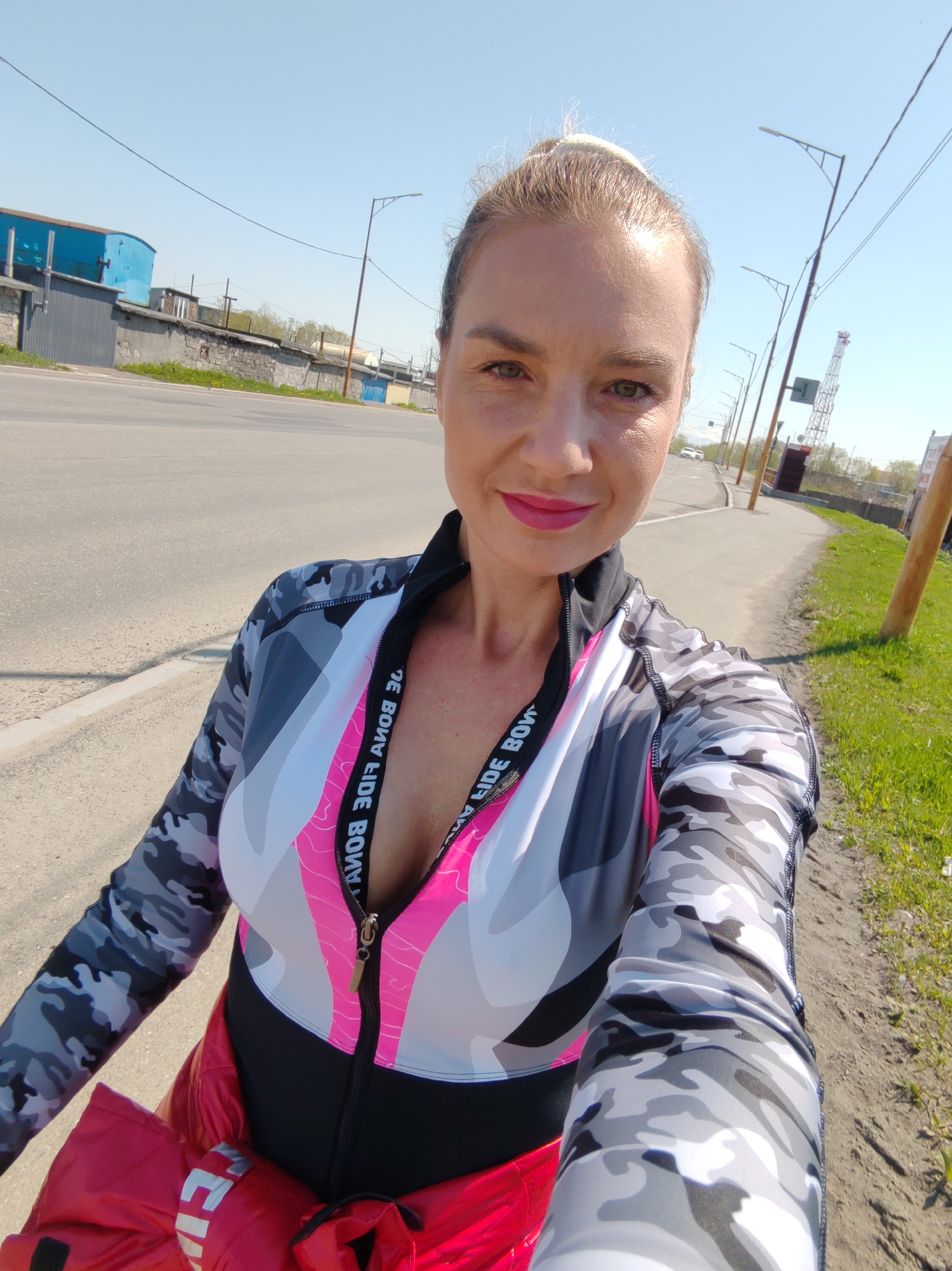 The image features a woman taking a selfie outdoors under a clear, bright blue sky. She is centered in the frame, directly facing the camera. She has blonde hair tied back in a bun, fair skin, green eyes, and is wearing pink lipstick. She is dressed in a long-sleeved athletic top with a gray, black, and white camouflage pattern on the sleeves and upper shoulders, and a geometric design in pink, white, and gray on the torso. The top has a partial zipper closure revealing a black underlayer, and mirrored text "BONA FIDE BONA" is visible on the inner trim. A bright red jacket is tied around her waist.
The background depicts a semi-urban environment. To the viewer's left, an asphalt road runs parallel to the foreground, separated by a narrow strip of dirt and sparse green grass from a series of buildings. These include a prominent bright blue building on the far left, followed by various gray, industrial or garage-like structures, some with stone-faced bases. Overhead, a black utility wire crosses the upper left portion of the sky.
To the viewer's right, a dirt path or sidewalk parallels the asphalt road, bordered by green grass and dirt. A series of modern streetlights with extended arms line the road, leading to a visible white car in the distance. Further back, a tall, red and white lattice telecommunication tower stands prominently. Wooden utility poles are visible along the right edge of the frame. Indistinct distant hills or mountains are visible under the clear sky on the horizon.
This scene, characterized by a mix of industrial-style buildings, asphalt roads, modern streetlights, a telecommunication tower, and the presence of distant hills or mountains, is consistent with peripheral or industrial districts within the city of Petropavlovsk-Kamchatsky, Russia. The terrain and infrastructure align with the typical landscape of this Far Eastern Russian city, situated amidst volcanic hills. A precise street location is not discernible from the provided imagery.