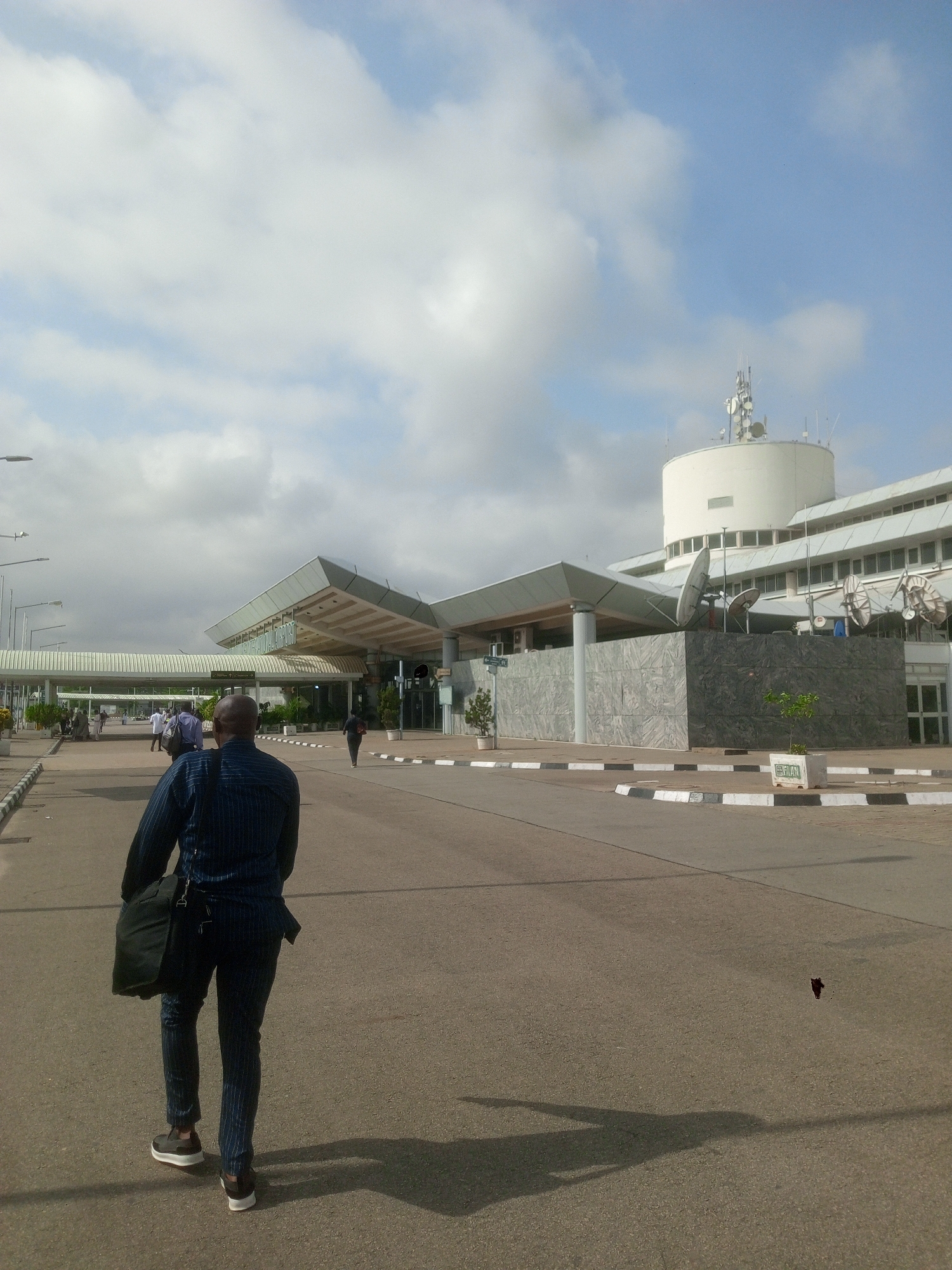 The image shows an individual walking away from the camera on a paved road in front of a building identified as "Unknown International Airport" in Nigeria. The sky is overcast with white clouds. Several other people are visible walking in the background near the airport entrance. The individual in the foreground is wearing a blue striped suit and carrying a black bag. The building has a distinctive cylindrical structure on top with multiple antennas and satellite dishes. The road is marked with black and white striped curbs. A small piece of debris is visible on the road surface.