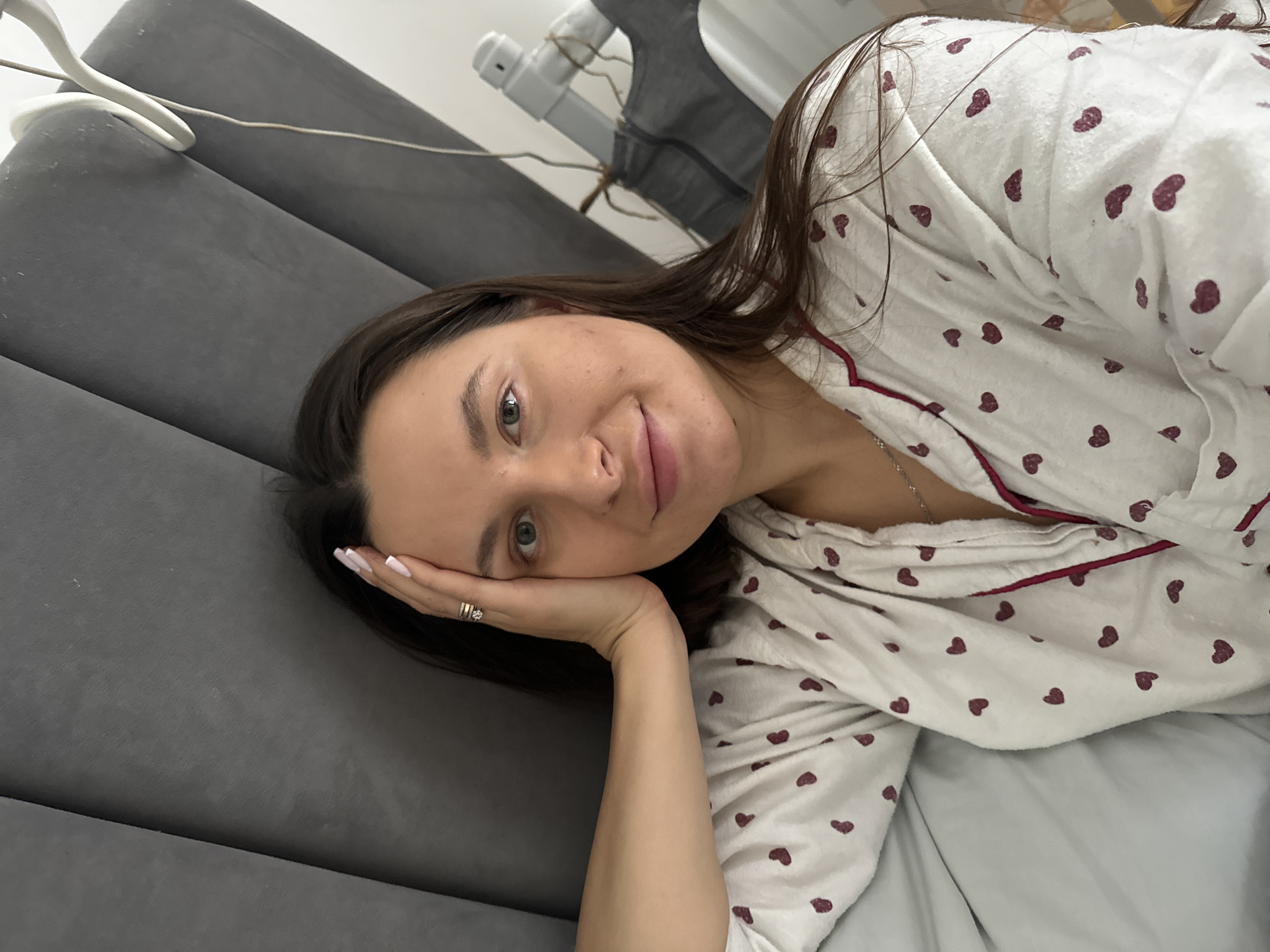 A woman with light skin and dark hair lies on a gray, padded surface, possibly a headboard or mattress, indoors. She is wearing a white pajama top adorned with small, maroon-colored hearts. Her left hand is supporting her head, and she has white nail polish on her nails. A gold ring is visible on her left ring finger. A portion of a hanger and what appears to be a clothing rack are visible in the background. The setting appears to be a bedroom or a similar domestic space. The location of the image within Akulovo (Gp Kubinka), Russia is not discernible from the visual information provided.