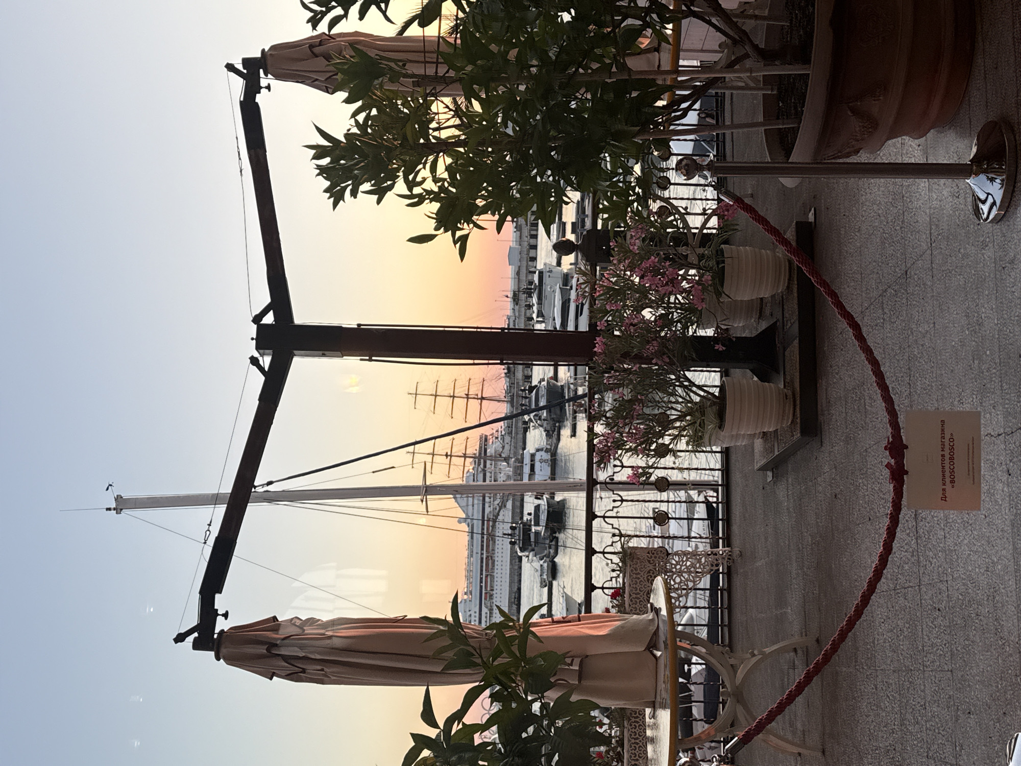 This photograph captures a serene sunset scene at a waterfront cafe in Sochi, Russia. The foreground features a paved terrace with several potted plants, including a large tree with green leaves and smaller plants with pink flowers. A red rope barrier with silver stanchions guides visitors, and a small sign in Russian is visible on the ground. In the midground, tables and chairs are set up, with one table partially visible in the left corner.
Beyond the terrace, a body of water, likely a harbor, is filled with various boats. A large white cruise ship is prominent in the distance, while several yachts and smaller vessels are closer to the shore. The water reflects the warm hues of the sunset, creating a shimmering effect. The sky is a gradient of soft orange, pink, and blue, typical of a sunset.
A large, dark metal structure with angled beams is positioned in the mid-foreground, supporting two large, folded beige umbrellas. The structure appears to be part of the cafe's outdoor seating area.
In the background, beyond the harbor, a distant shoreline with buildings is visible. The overall atmosphere is peaceful and picturesque, highlighting the beauty of a coastal city at dusk. The reflections in the water and the soft lighting contribute to the tranquil mood.